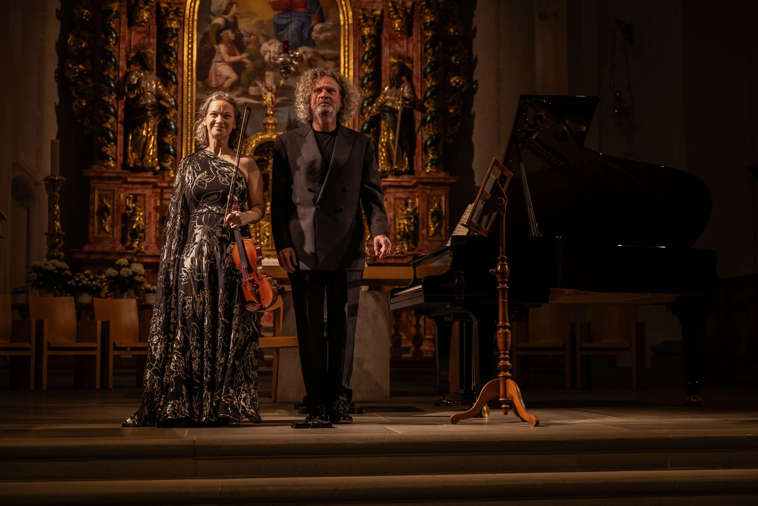 Two musicians, a woman holding a violin and a man standing beside her, on a stage in a church or cathedral with religious artwork in the background, a grand piano nearby, after a performance.
