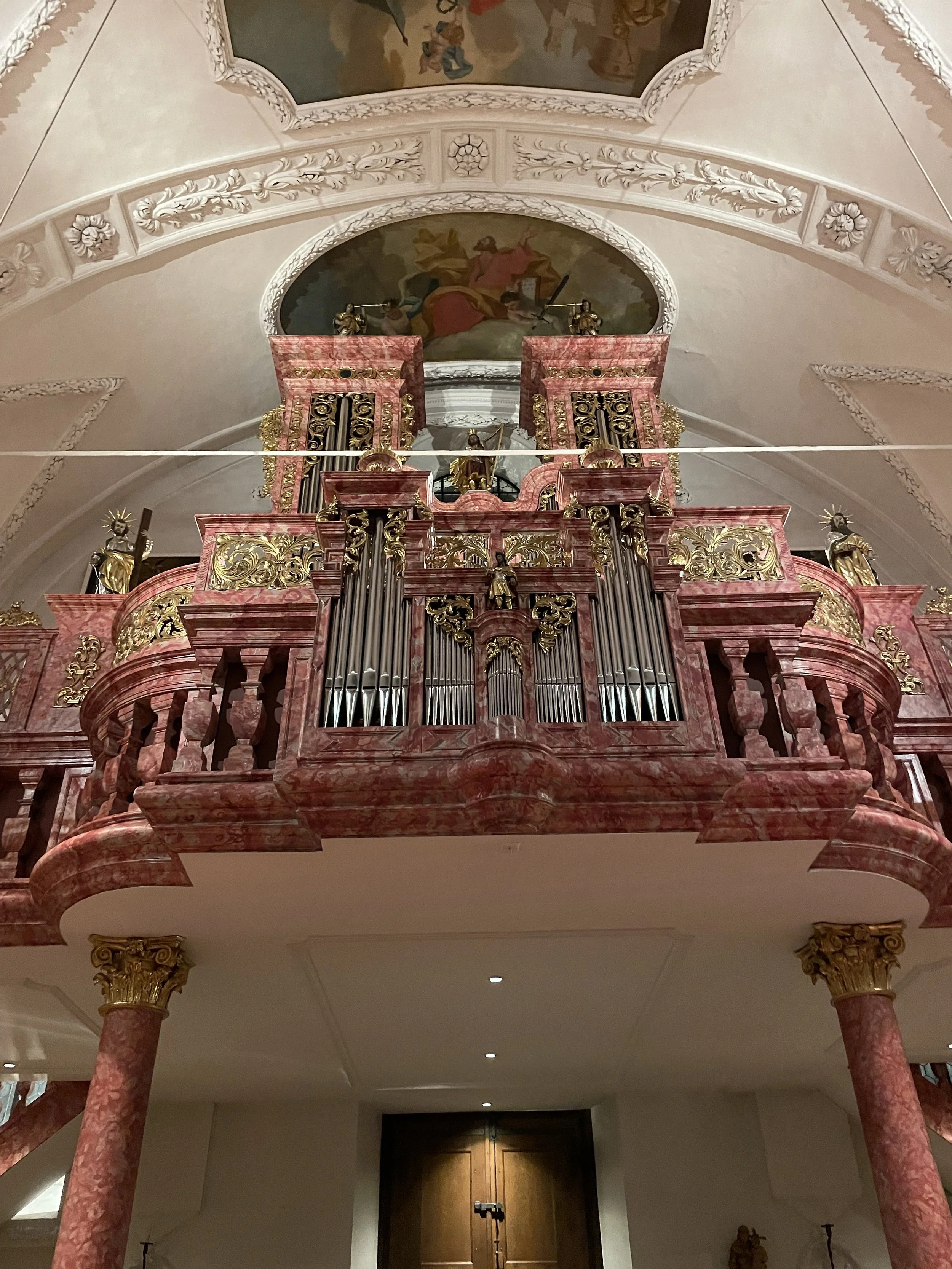 A church interior with a large ornate pink and gold pipe organ on a balcony, supported by pink marble columns with gold accents. Above the organ, there are religious statues on the balcony and a ceiling painting depicting a biblical scene.