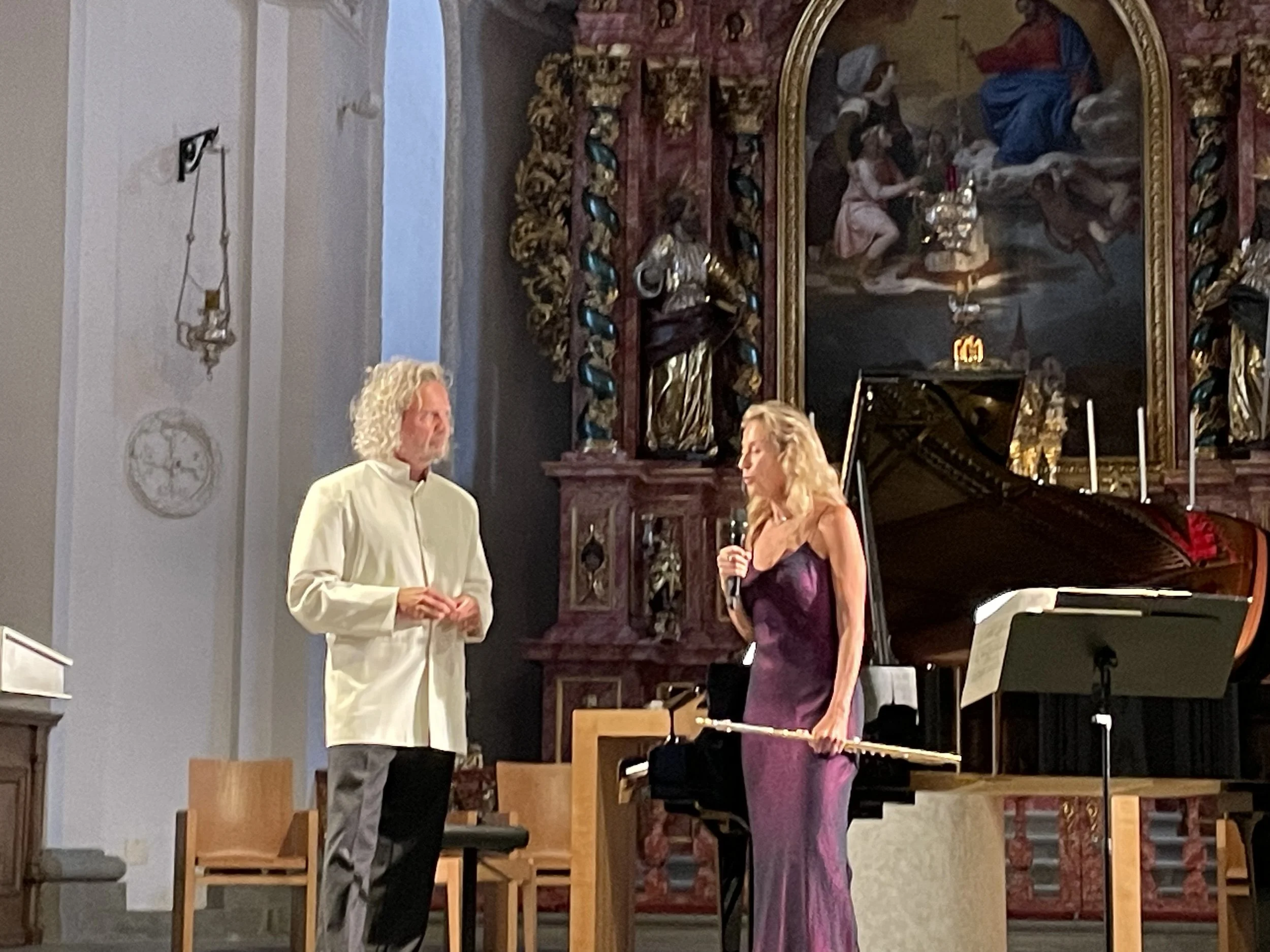 A woman in a purple dress holding a flute and a man in a white coat standing on a stage inside a church or concert hall, with a grand piano and ornate religious art in the background.