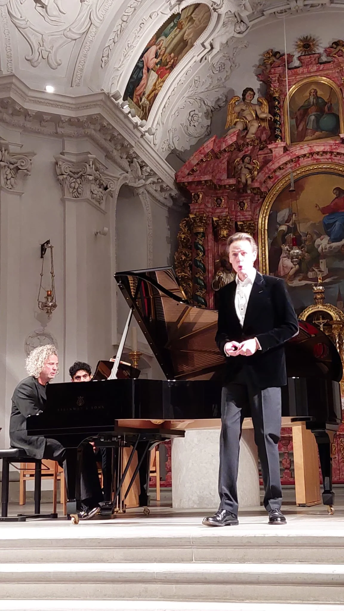 Three musicians performing on a stage inside a church or cathedral with ornate decorations and religious paintings. One person is playing a grand piano, another is standing and singing or speaking, and the third is partially visible behind the piano.