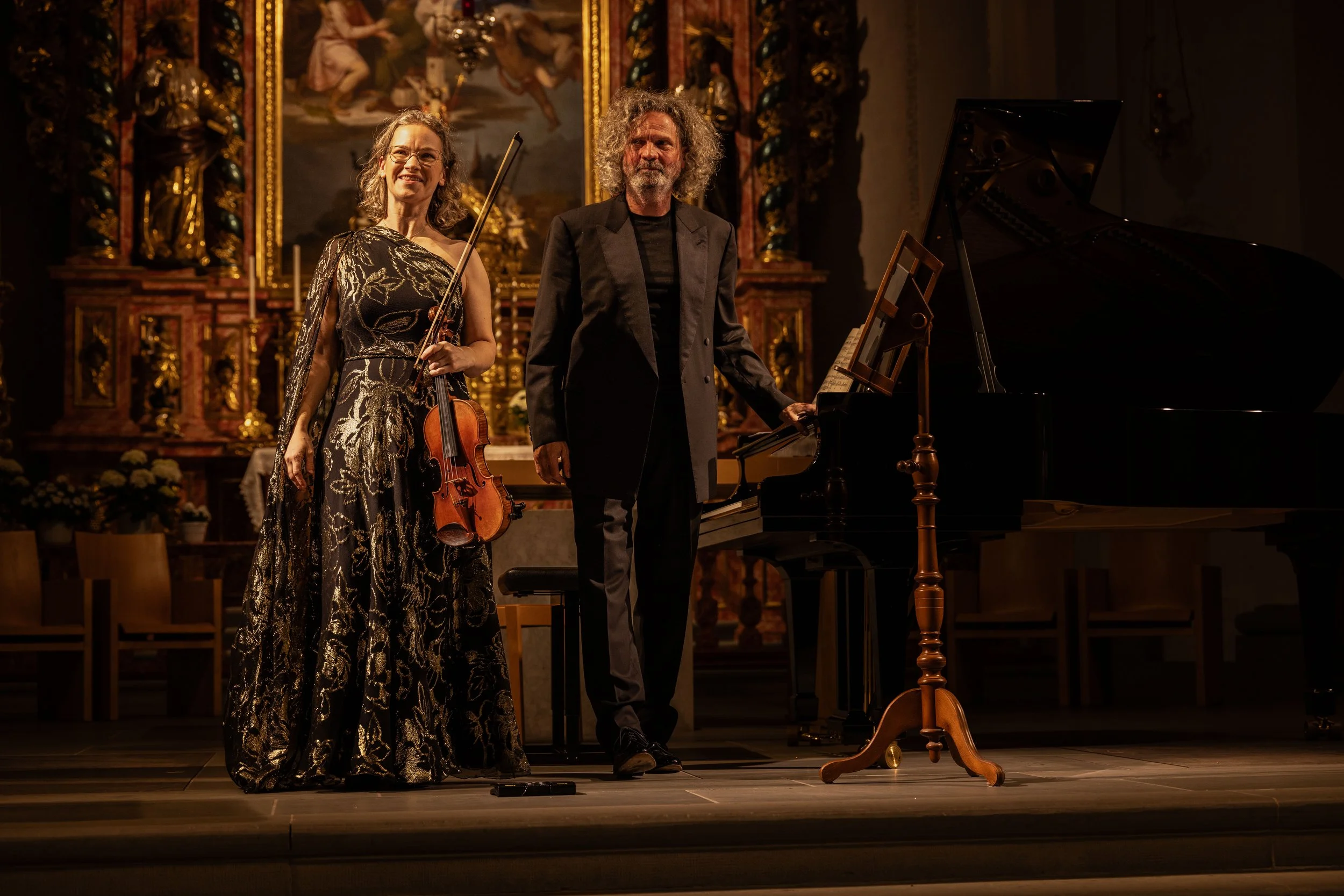 Two musicians, a woman with glasses holding a violin and bow, and a man with curly hair standing next to a grand piano, take a bow on stage in a decorated concert hall.