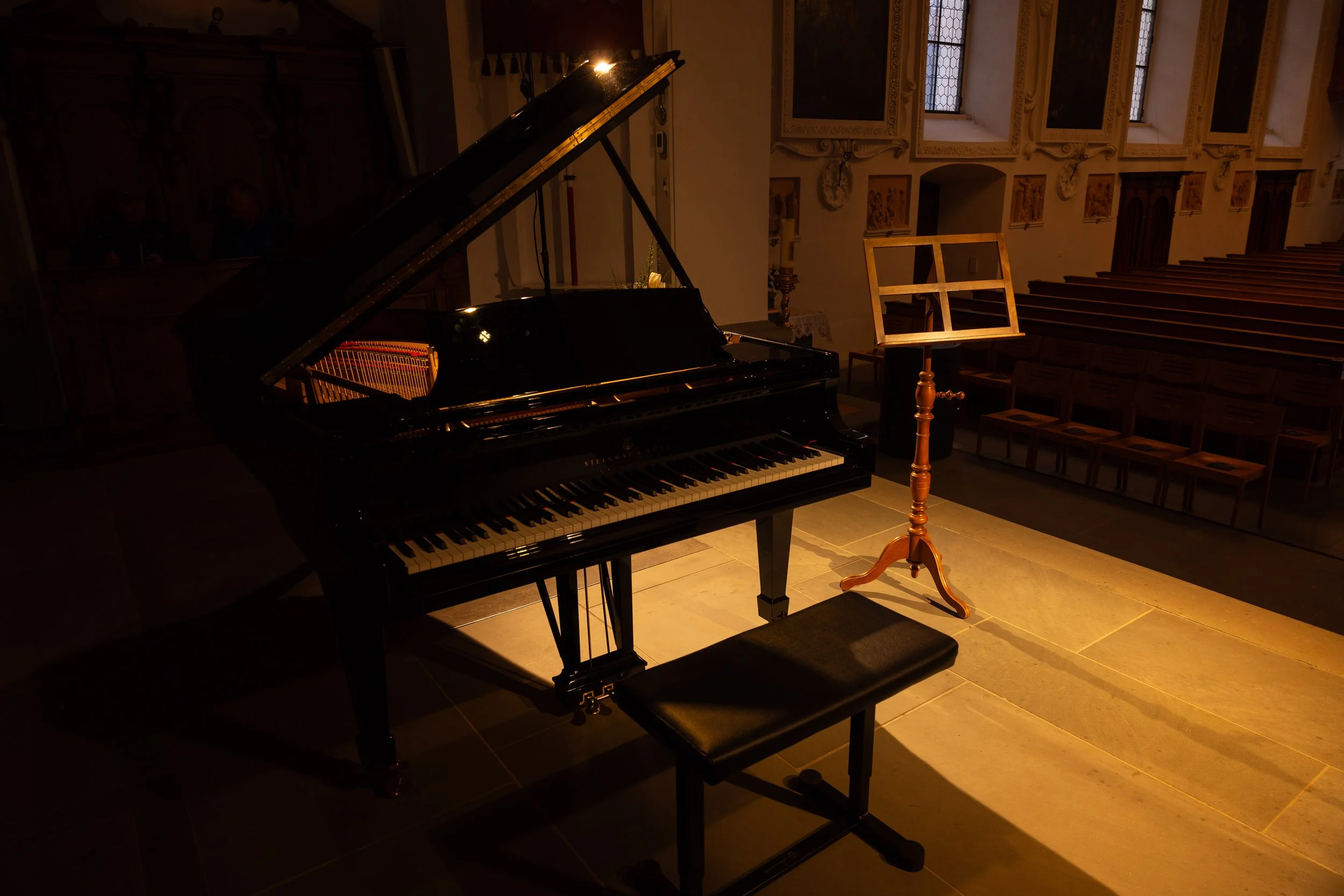 A grand piano with an open lid, set in a dimly lit church or concert hall, with a wooden music stand and a matching bench nearby, and rows of wooden pews in the background.