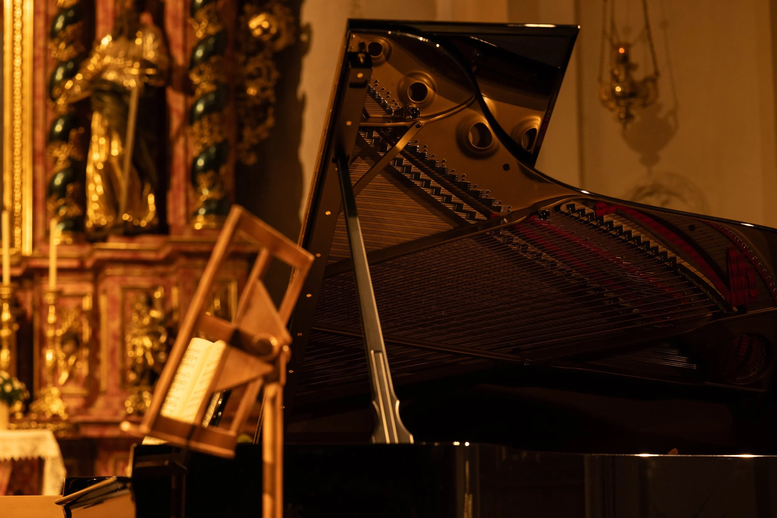 A black grand piano with its lid open, piano strings and hammers visible, set in a dimly lit room with ornate decorations in the background.