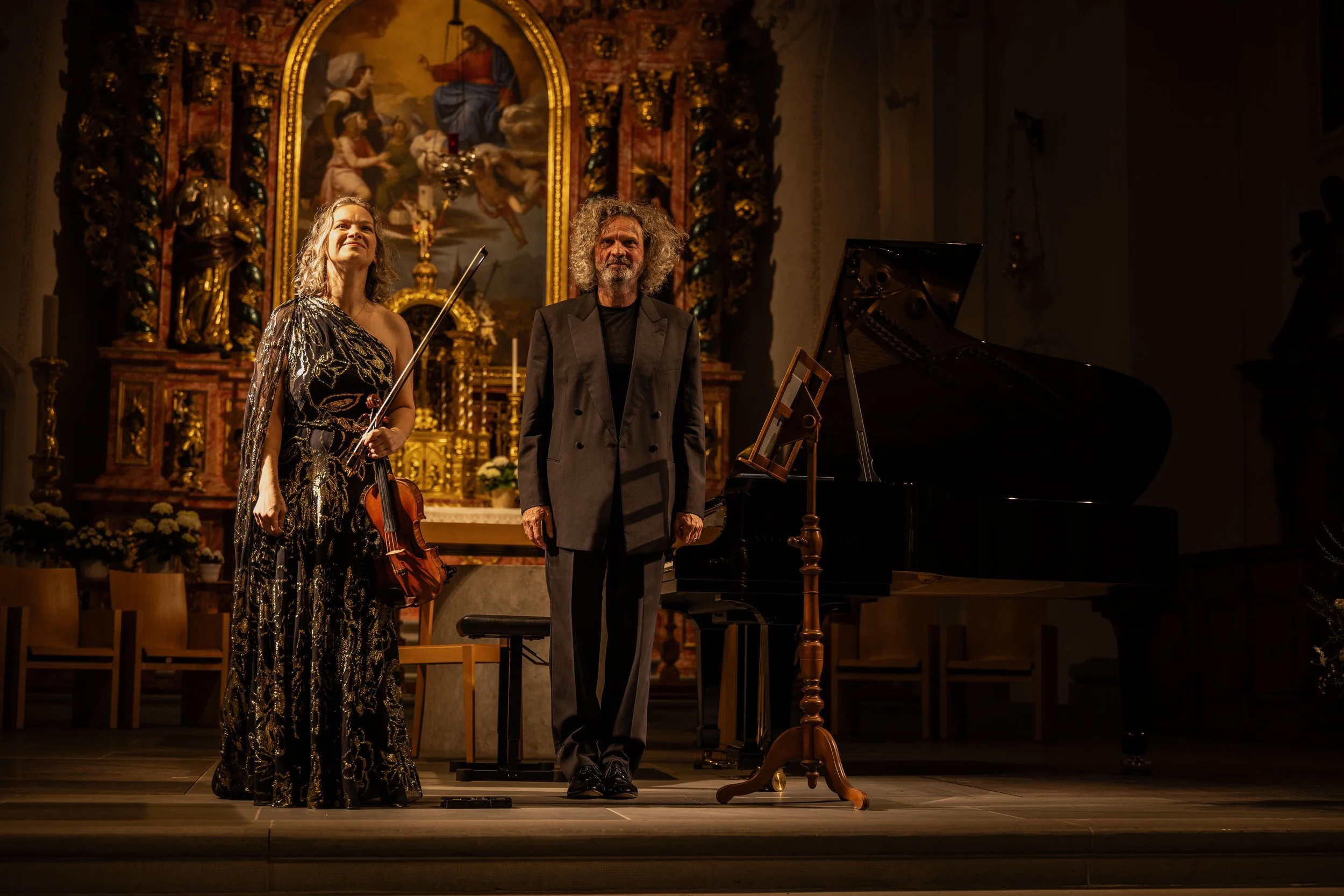 Two performers, a woman holding a violin and a man, standing on a stage in a church or church-like setting with a grand piano and an ornate altar with religious artwork and statues in the background.