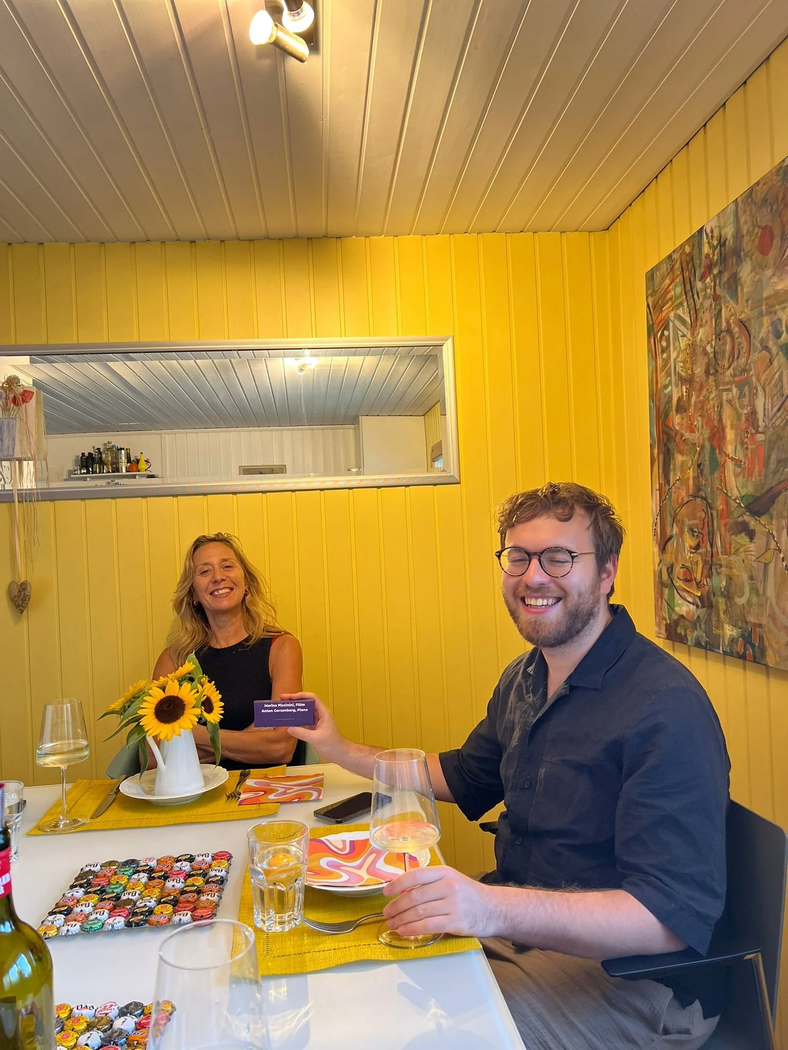 A man and a woman celebrating at a dinner table with flowers and drinks. The man is smiling and handing a purple card to the woman, who is also smiling. The room has yellow walls and colorful artwork, with a mirror on the wall reflecting the ceiling