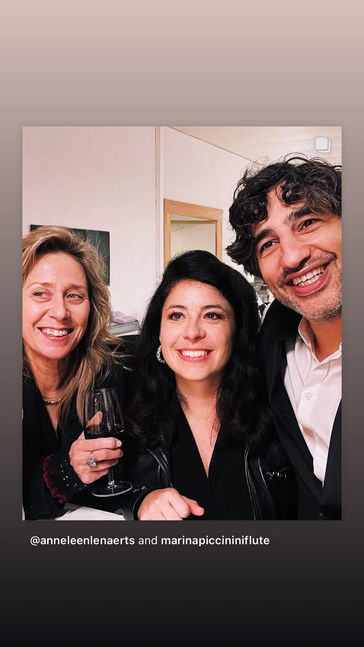 Three people smiling and posing for a photo at a social gathering, with one holding a glass of red wine.