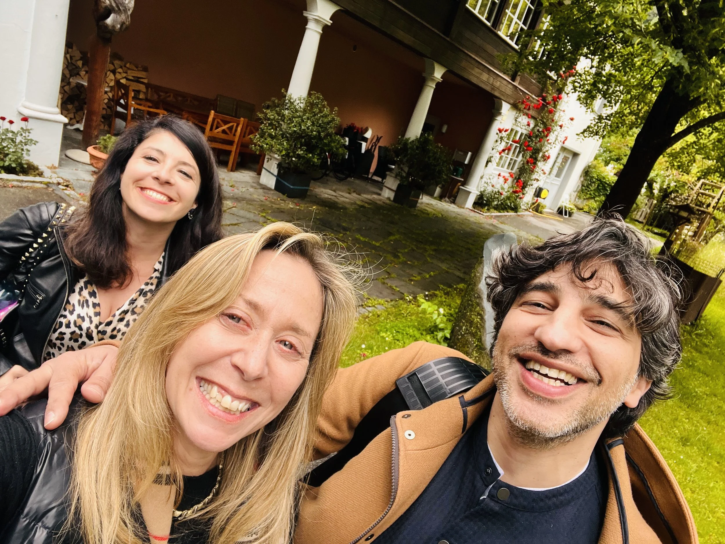 A group of three people taking a selfie outdoors in a garden or yard, smiling at the camera with a house and trees in the background.
