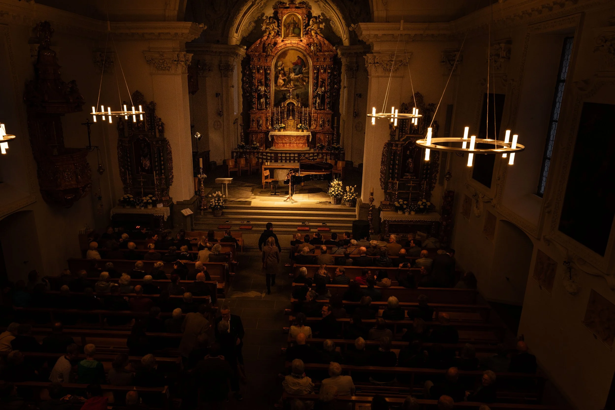 A church interior during a concert with an audience seated on pews, a grand piano on the stage, and ornate decorations and candles illuminating the space.