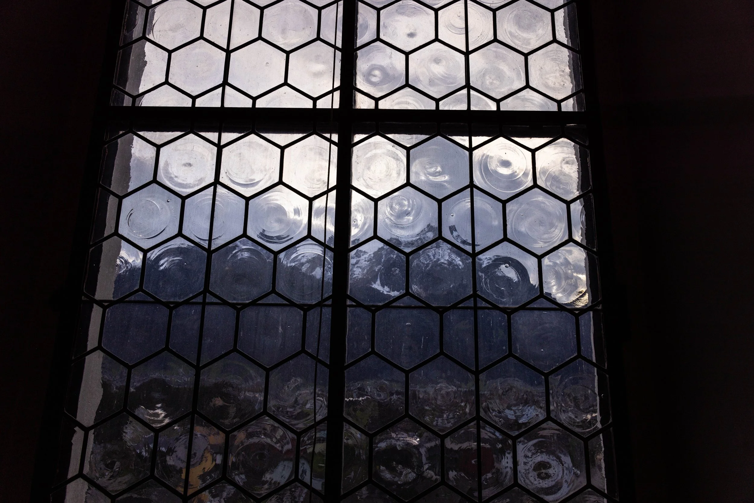 View through a stained glass window with hexagonal glass panes, showing a cloudy sky and distant mountains outside.