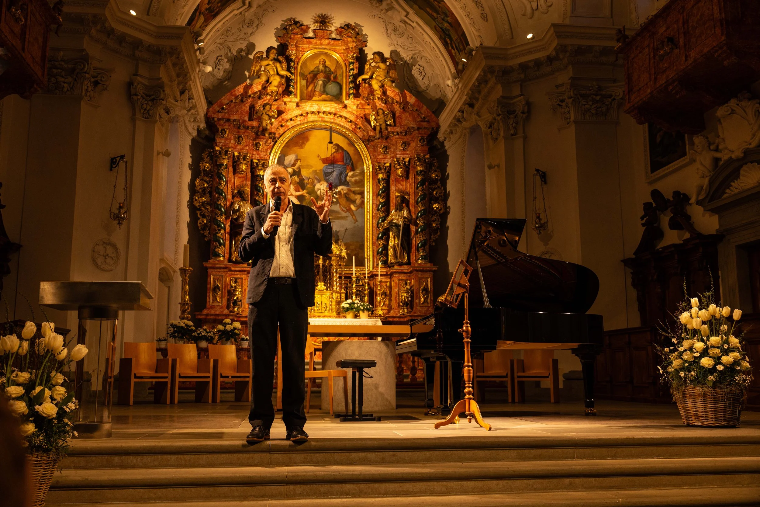 A man holding a microphone speaking on a stage inside a church with an ornate altar in the background. There is a grand piano and floral arrangements on either side of the stage.