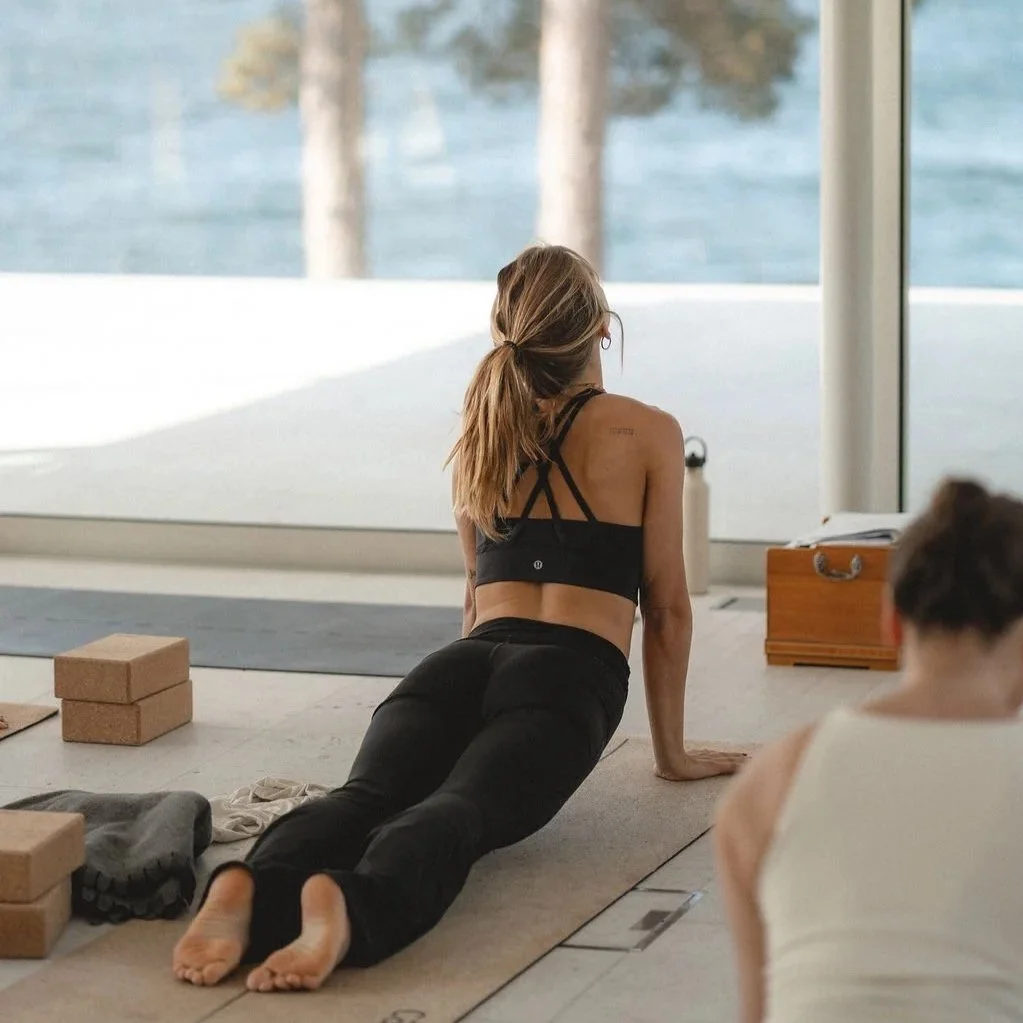 A woman in black athletic wear doing a yoga pose on a mat indoors near large windows overlooking a body of water and trees.