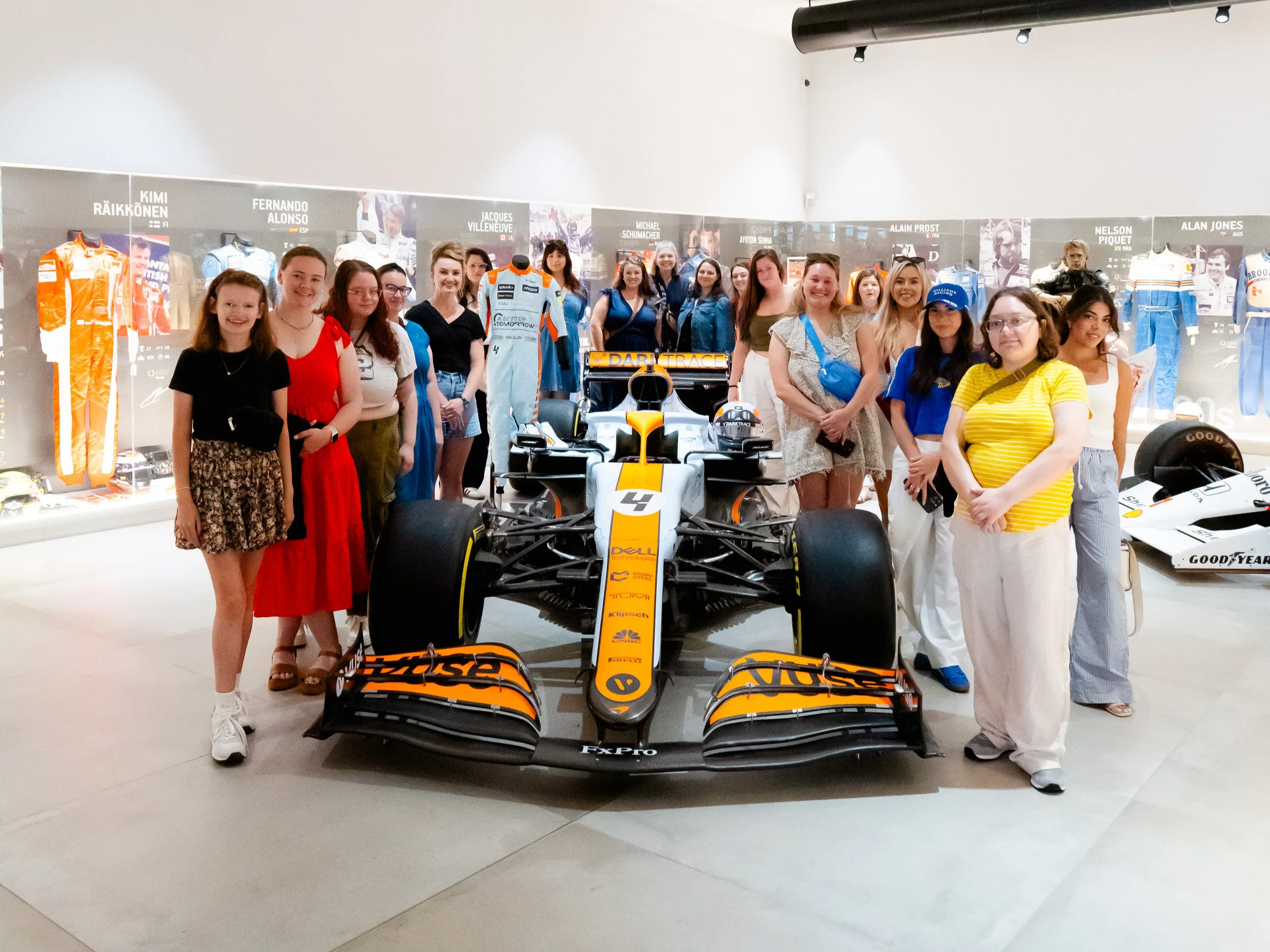 OTTR guests posing with Lando Norris's McLaren F1 car during a private tour of a racing car collection with historic driver suits and memorabilia on display