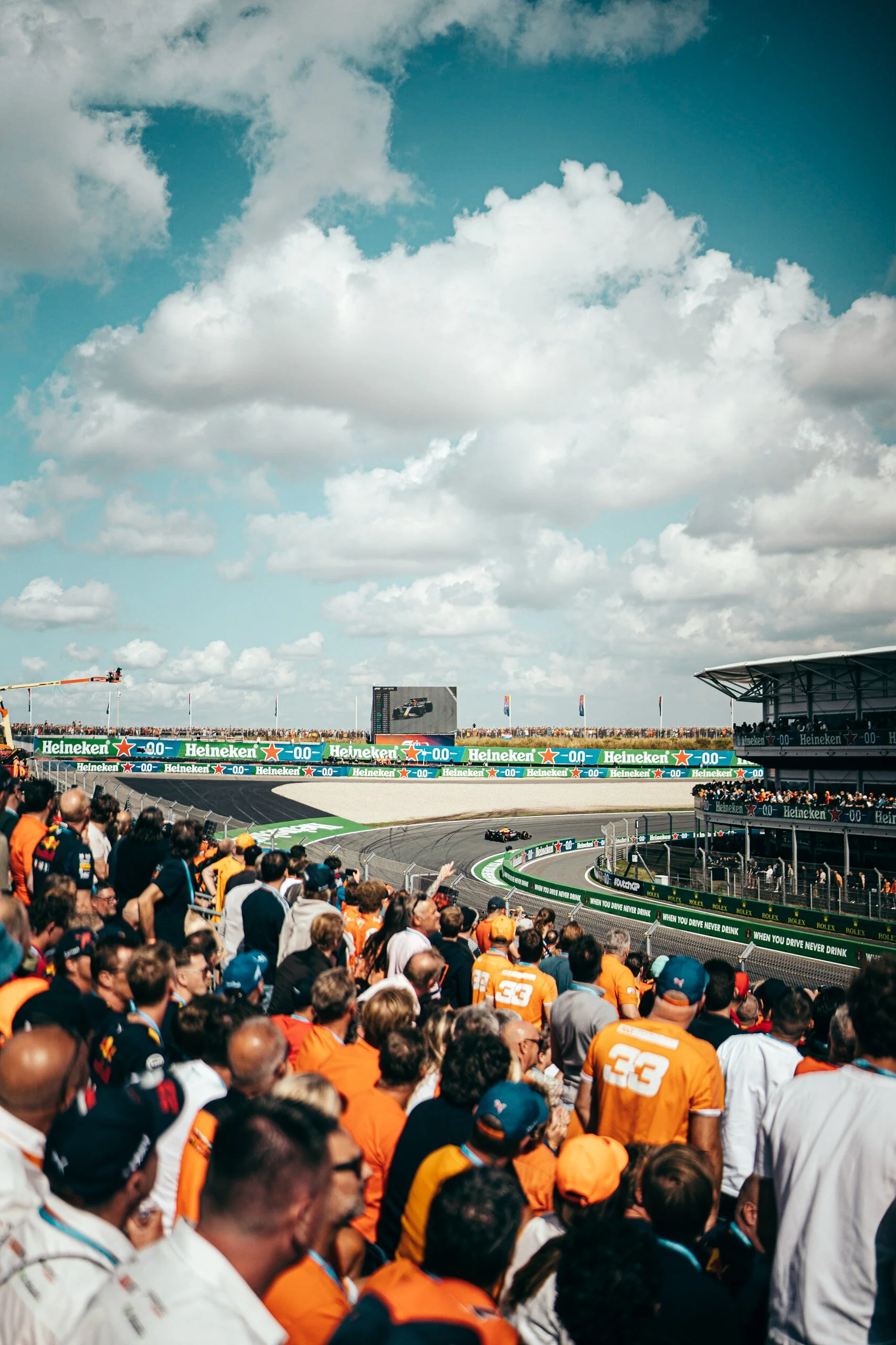 View from the grandstands at Tarzan corner (Turn 1) at Circuit Zandvoort during the Dutch Grand Prix, with F1 cars racing through the banked hairpin and fans in orange filling the stands under a wide blue sky