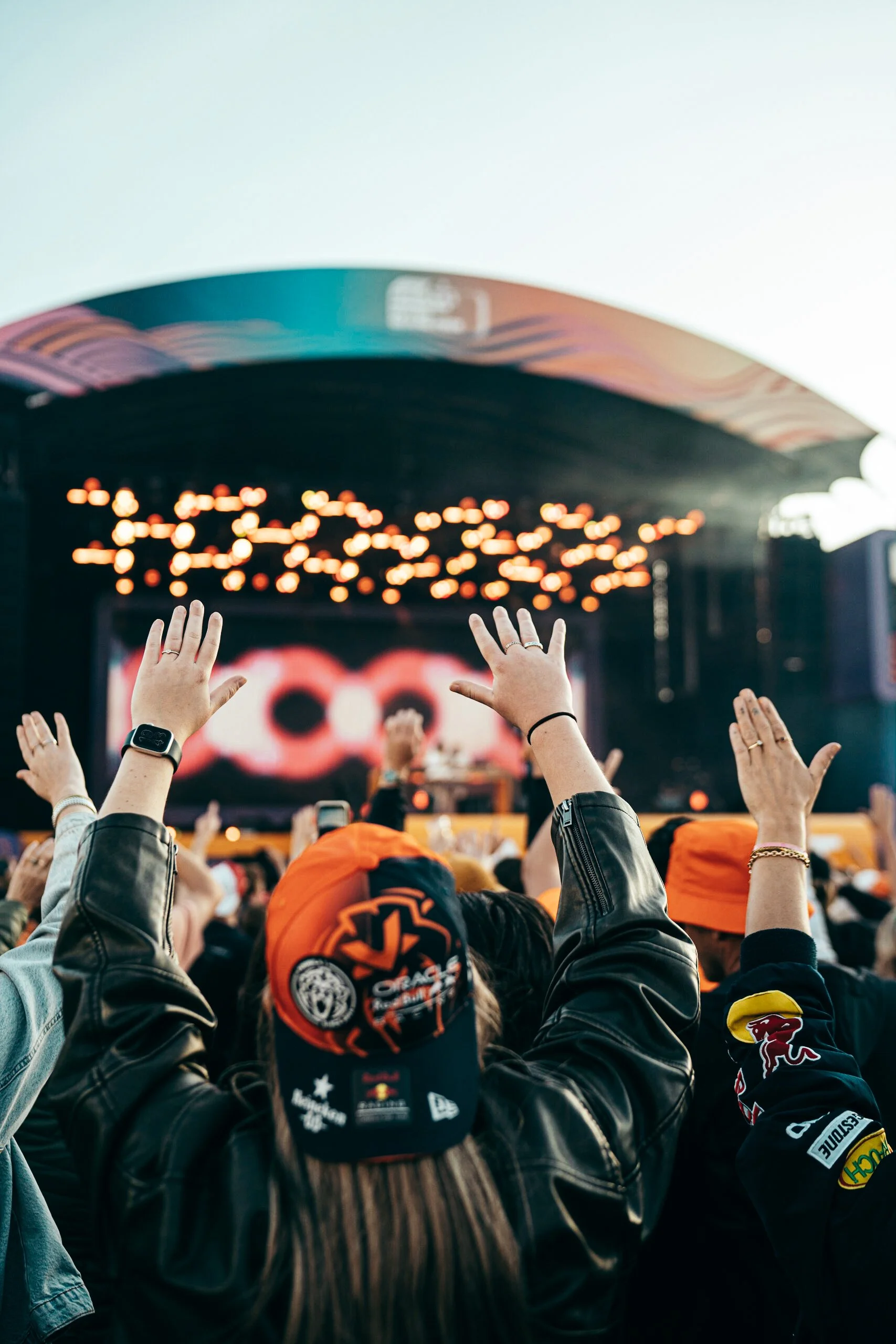 Fans with hands raised at a Dutch Grand Prix concert in the Zandvoort Fanzone, wearing Red Bull Racing and Max Verstappen gear with stage lights glowing in the background