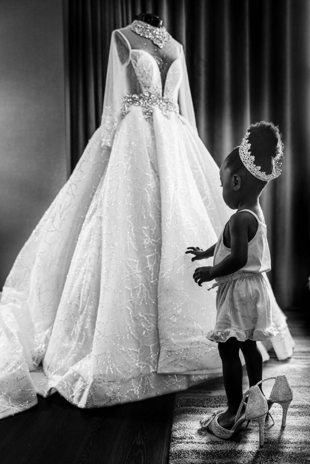 A-flower-girl-standing-in-the-brides-high-heels-admiring-her-wedding-dress.-Black-and-white-image-with-beautiful-lighting.-.jpeg