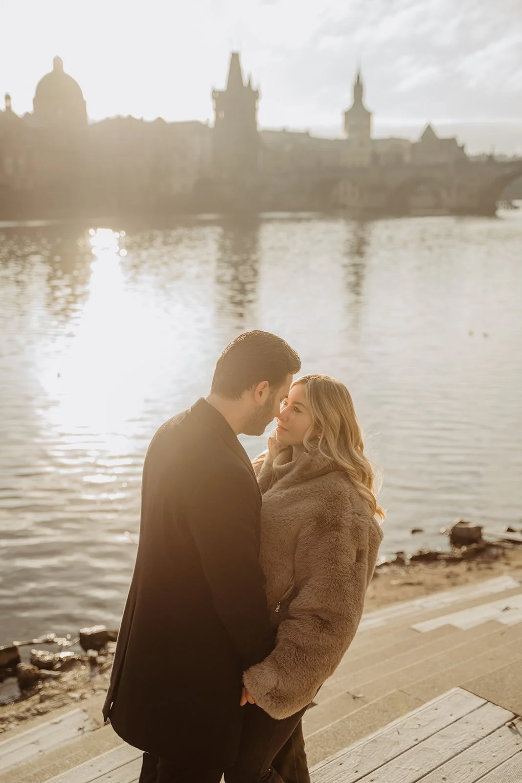 A couple stands close together on a wooden dock near a body of water, with a city skyline in the background, during sunset.