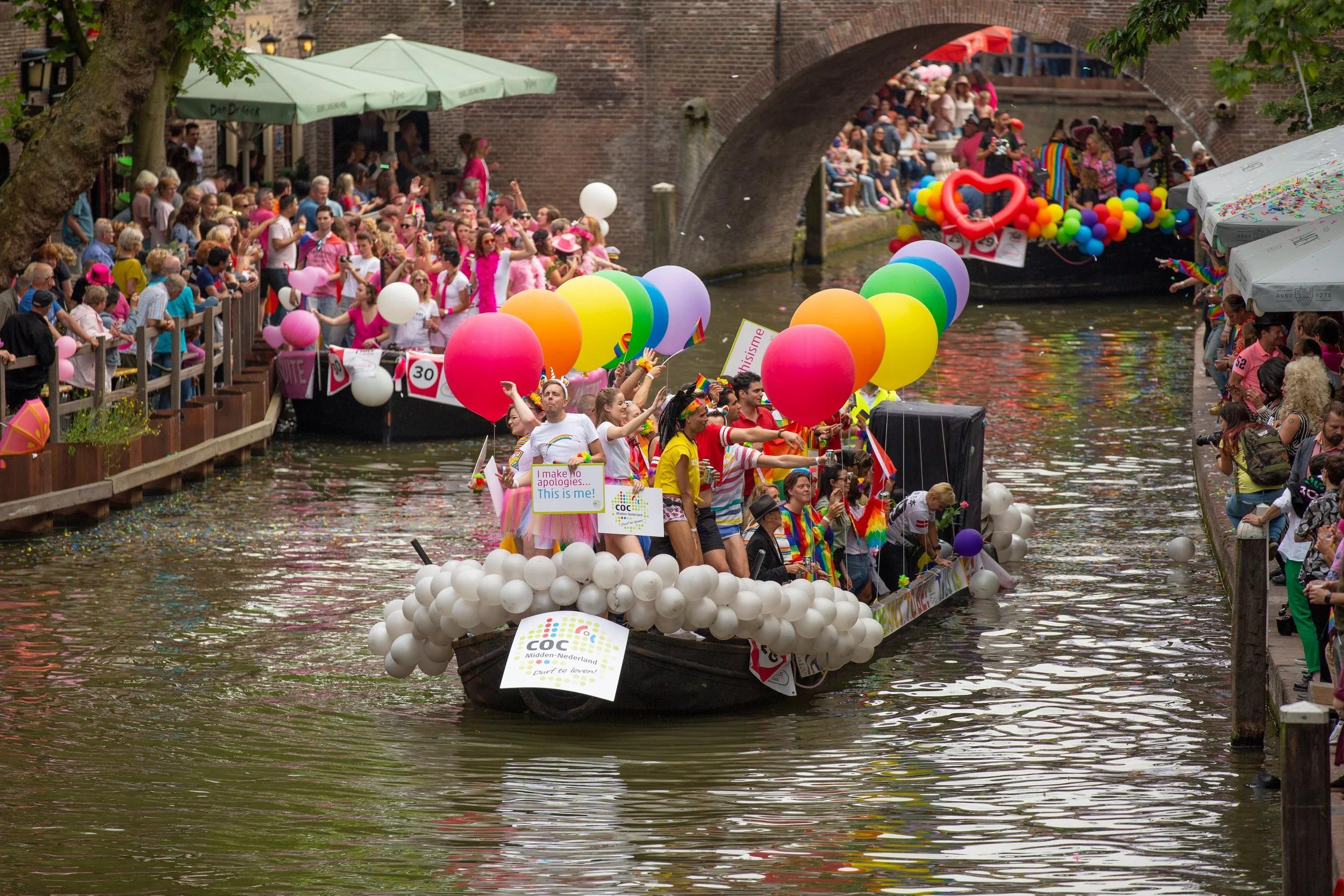Pride parade boats on a canal with rainbow balloons, flags and crowds celebrating LGBTQ+ visibility, inclusion and community belonging during Pride Month celebration.