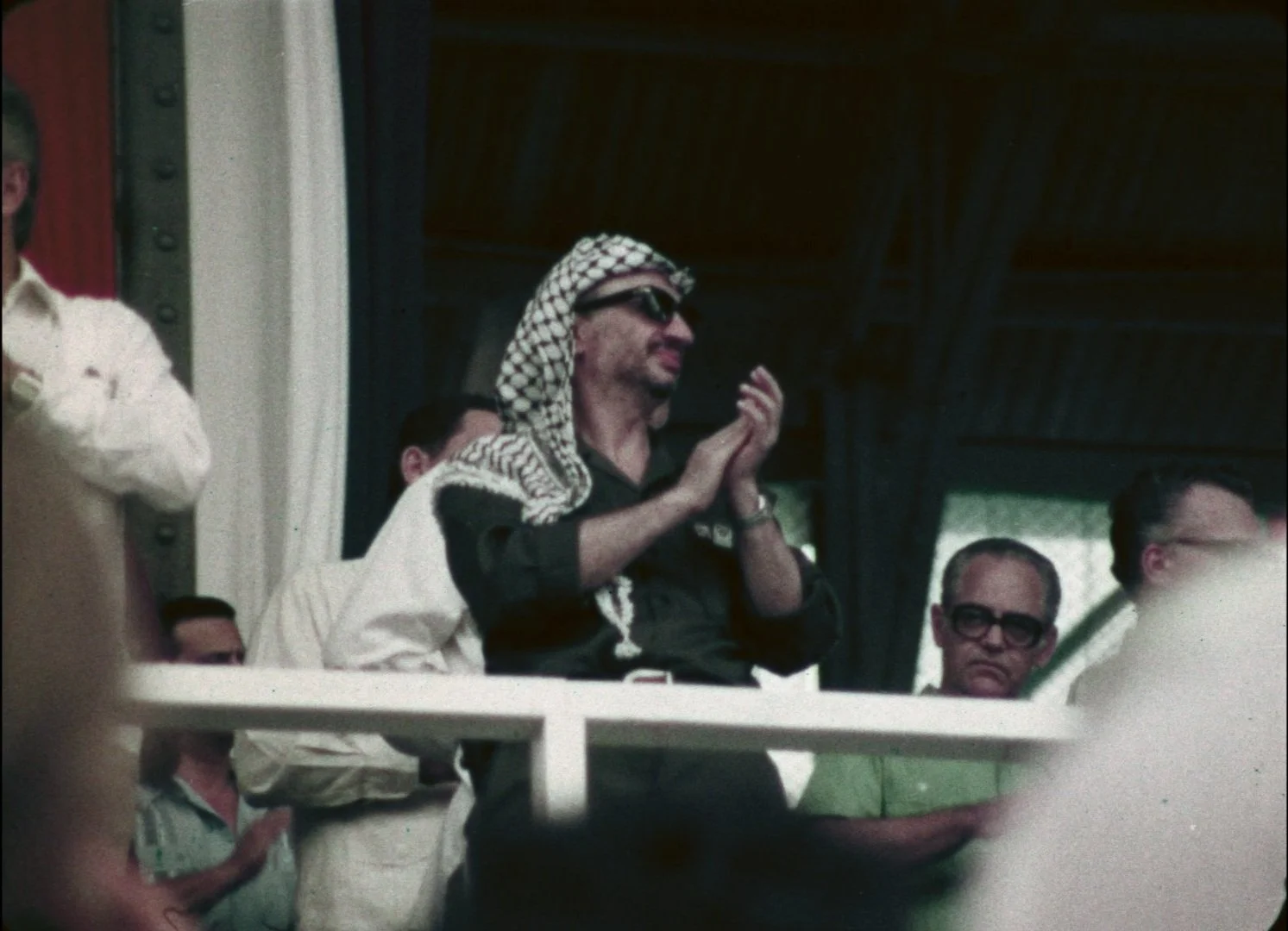 Yasser Arafat wearing sunglasses and a keffiyeh headscarf clapping among a group of men at an outdoor event.