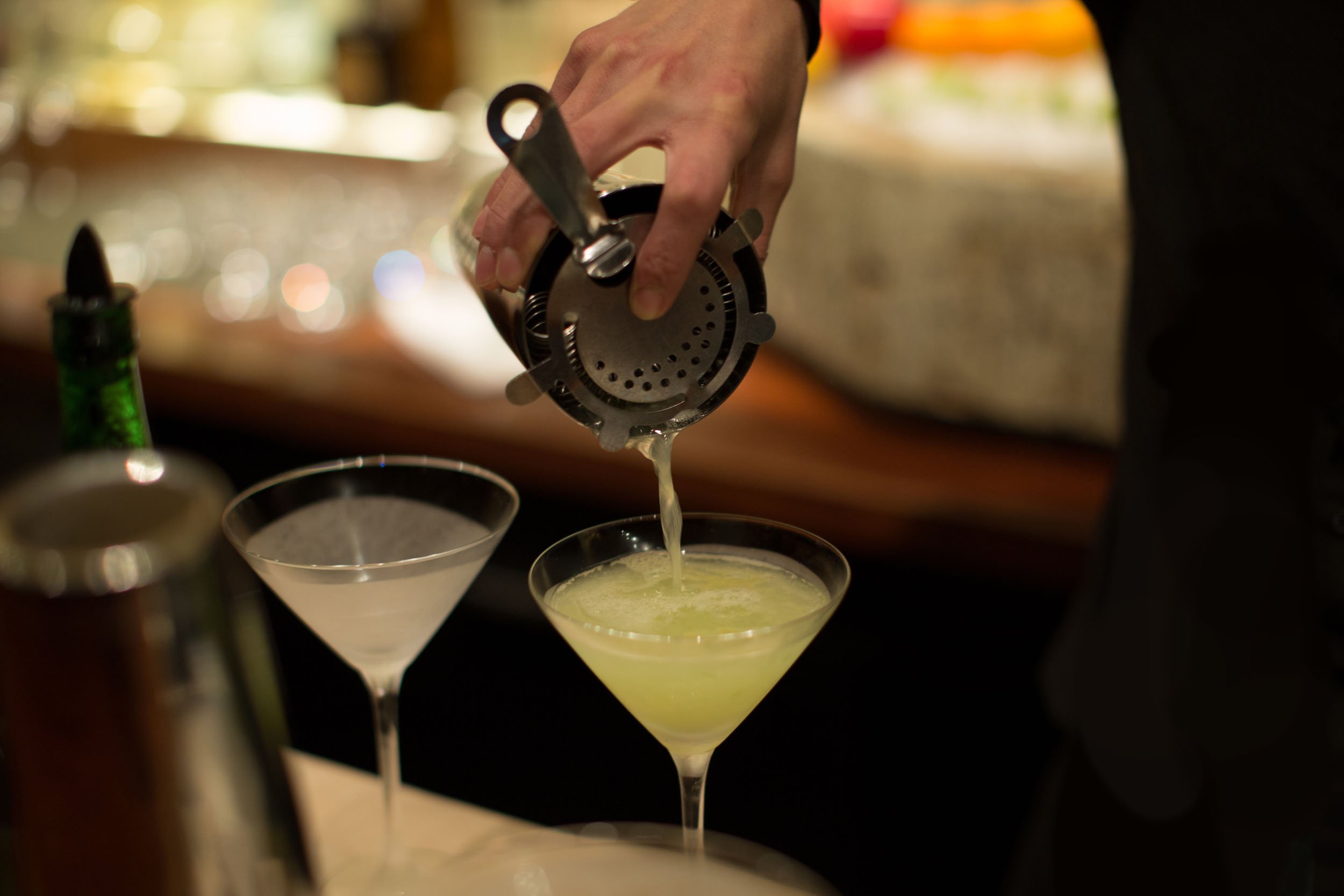  Closeup of a bartender’s hand pouring a green cocktail from a shaker into a martini glass, which is next to another filled glass. 