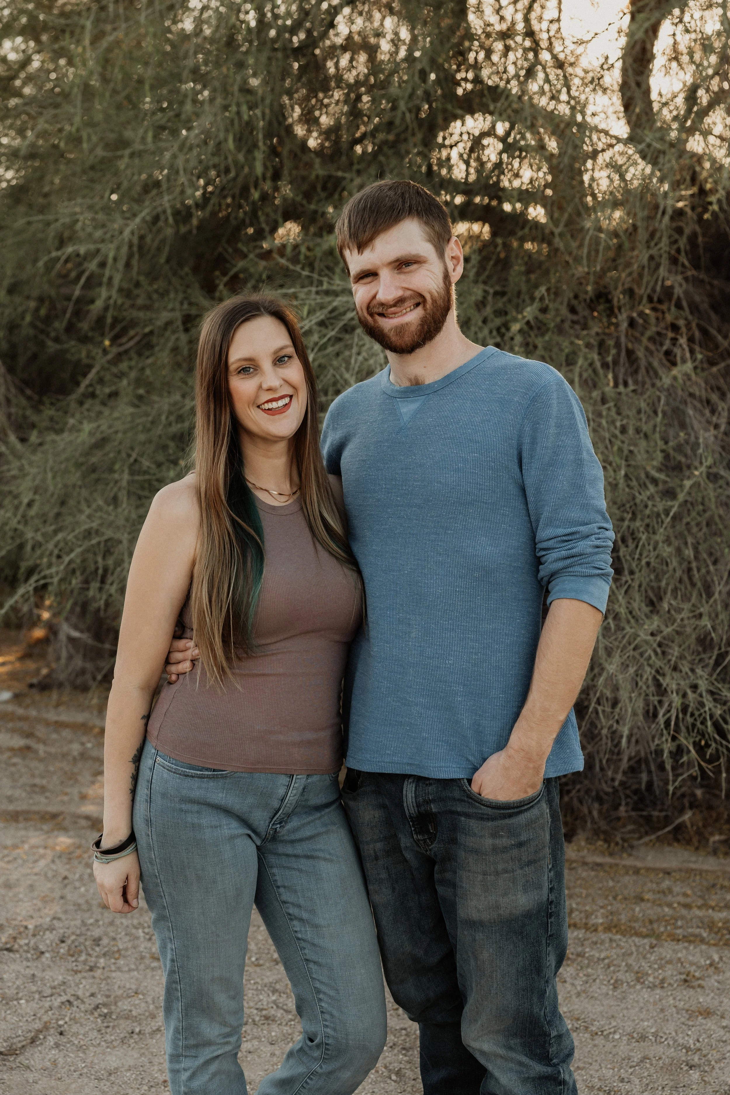 A smiling couple posing outdoors in front of trees during sunset, with the woman on the left and the man on the right.