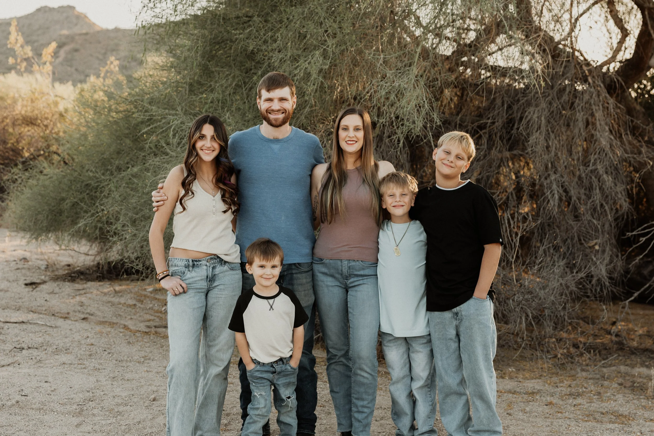 A group of six people, including three children and three adults, standing outdoors in front of trees and bushes during daytime.