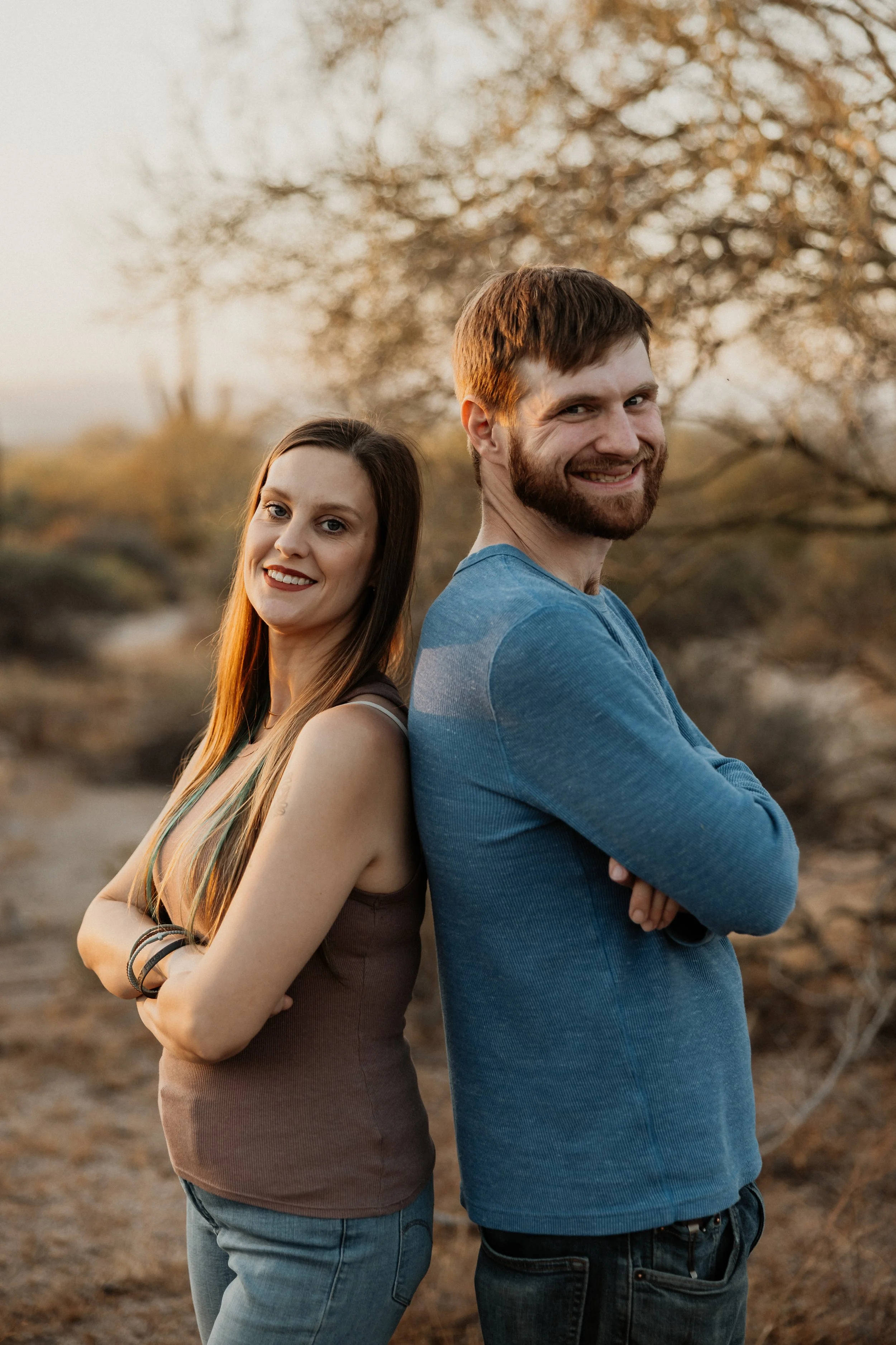 A man and woman stand back-to-back outdoors with arms crossed, smiling at the camera during sunset or late afternoon, with blurred trees and natural background.