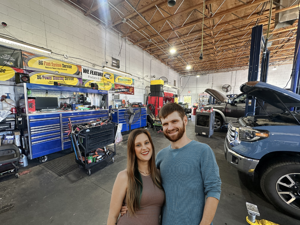 A smiling woman and man standing inside an automotive repair shop with car repair tools, blue tool chests, and vehicles with open hoods in the background.