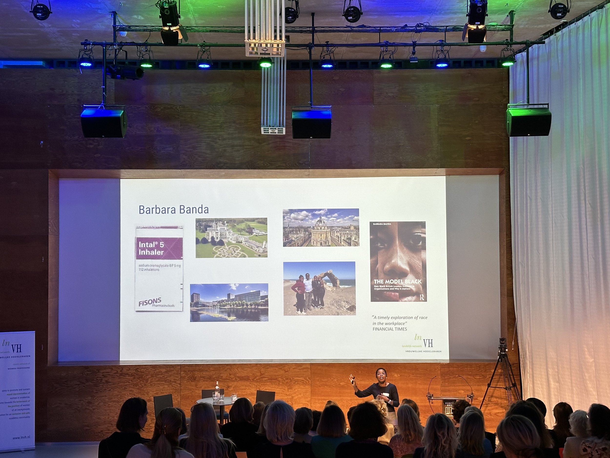 A woman giving a presentation in a large auditorium with a screen behind her displaying images and text about Barbara Banda, including pictures of buildings and a book cover, with an audience seated and watching.