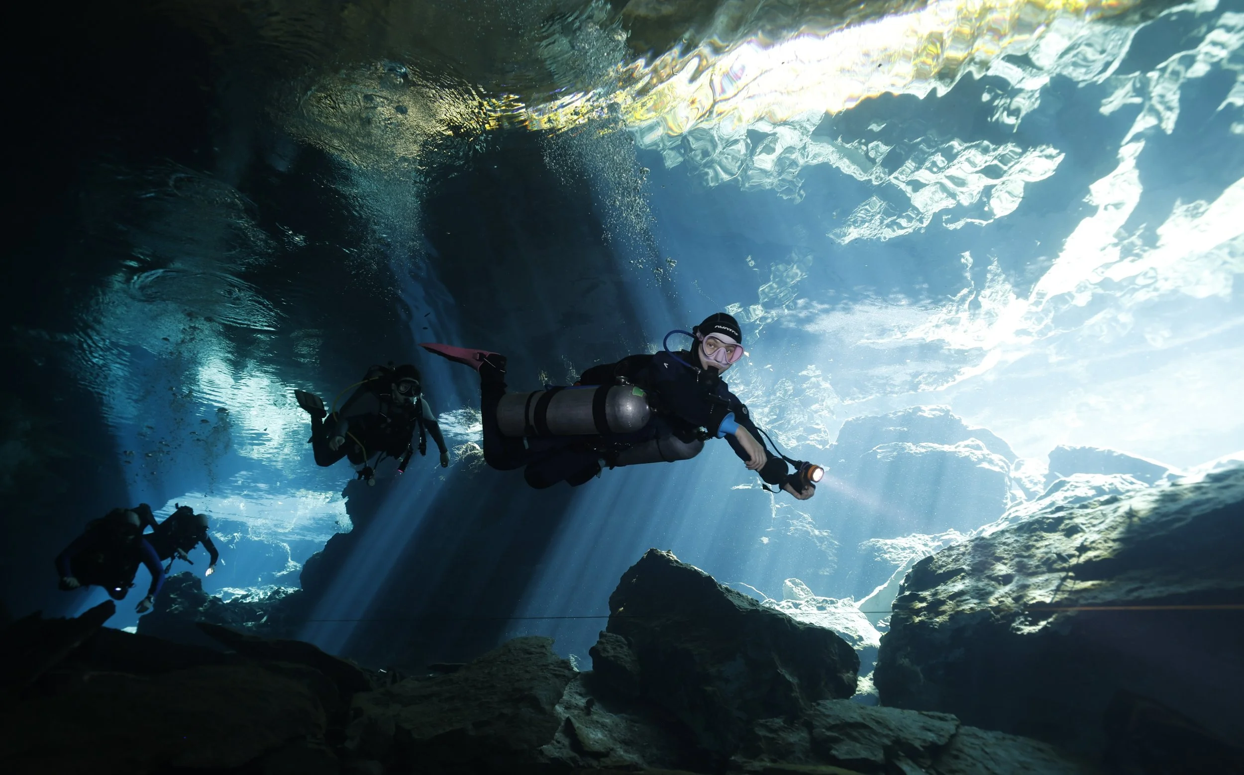 Divers exploring an underwater cave with sunlight streaming through the water.
