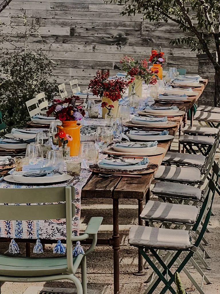 Outdoor dining table set with plates, glasses, and colorful flower centerpieces on a long wooden and fabric table, surrounded by chairs with cushions, against a weathered wooden fence and nearby trees.