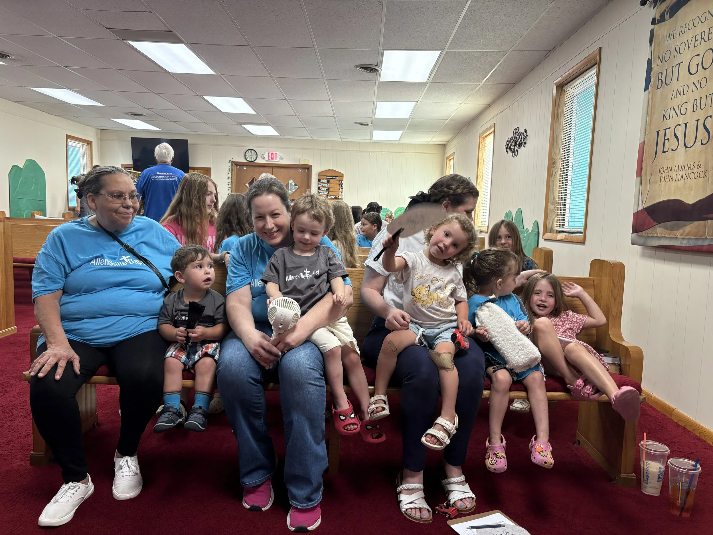 Children and adults sitting on pews inside a church. Everyone smiling and enjoying their time. Some children are holding items like a fan and a toy sword. Colorful wall decor and a large banner with religious text are visible.