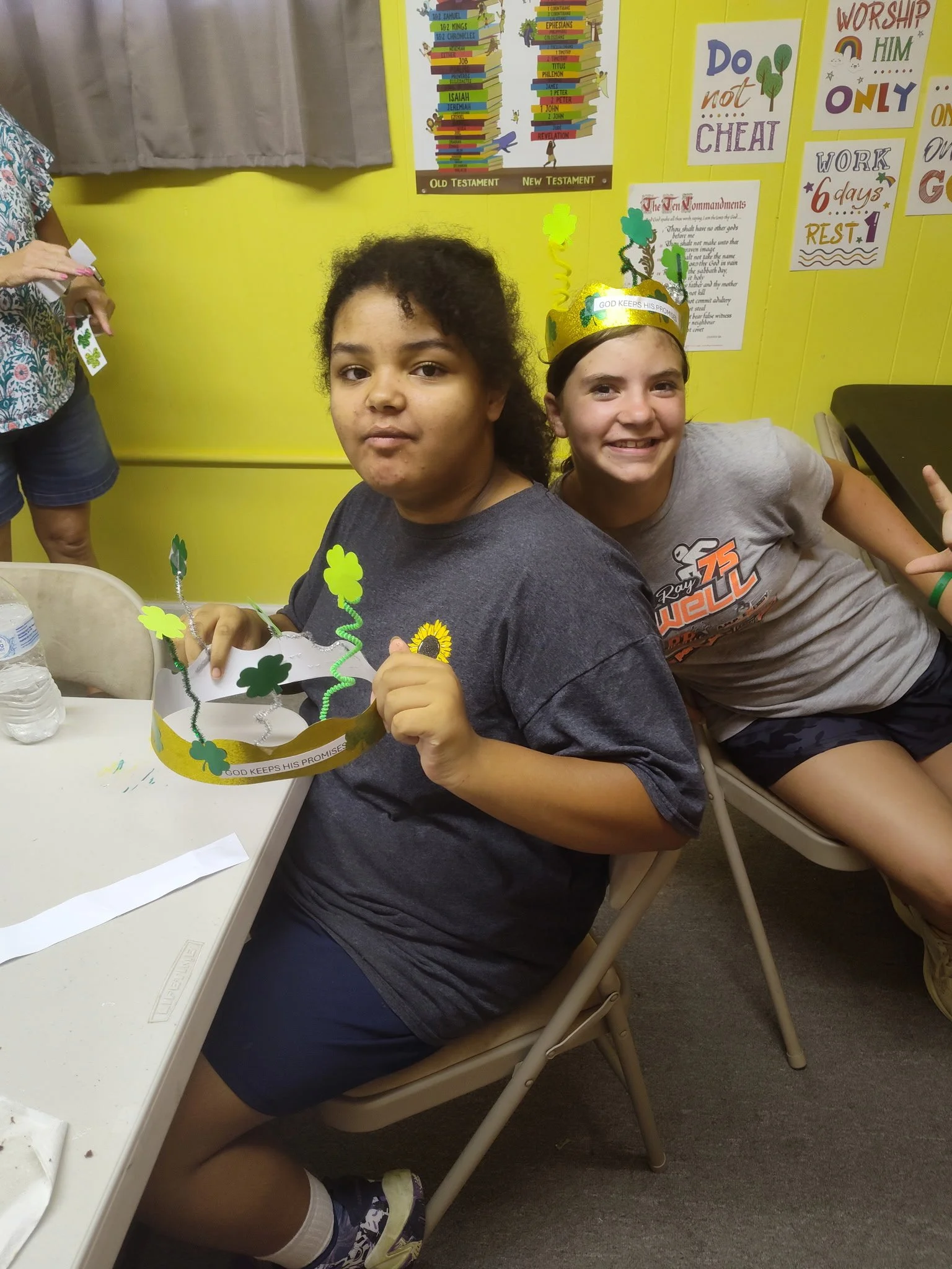 Two girls sitting at a table in a classroom, wearing paper crowns decorated with shamrocks and spring-themed items, smiling and making gestures. The background features colorful posters and a yellow wall.
