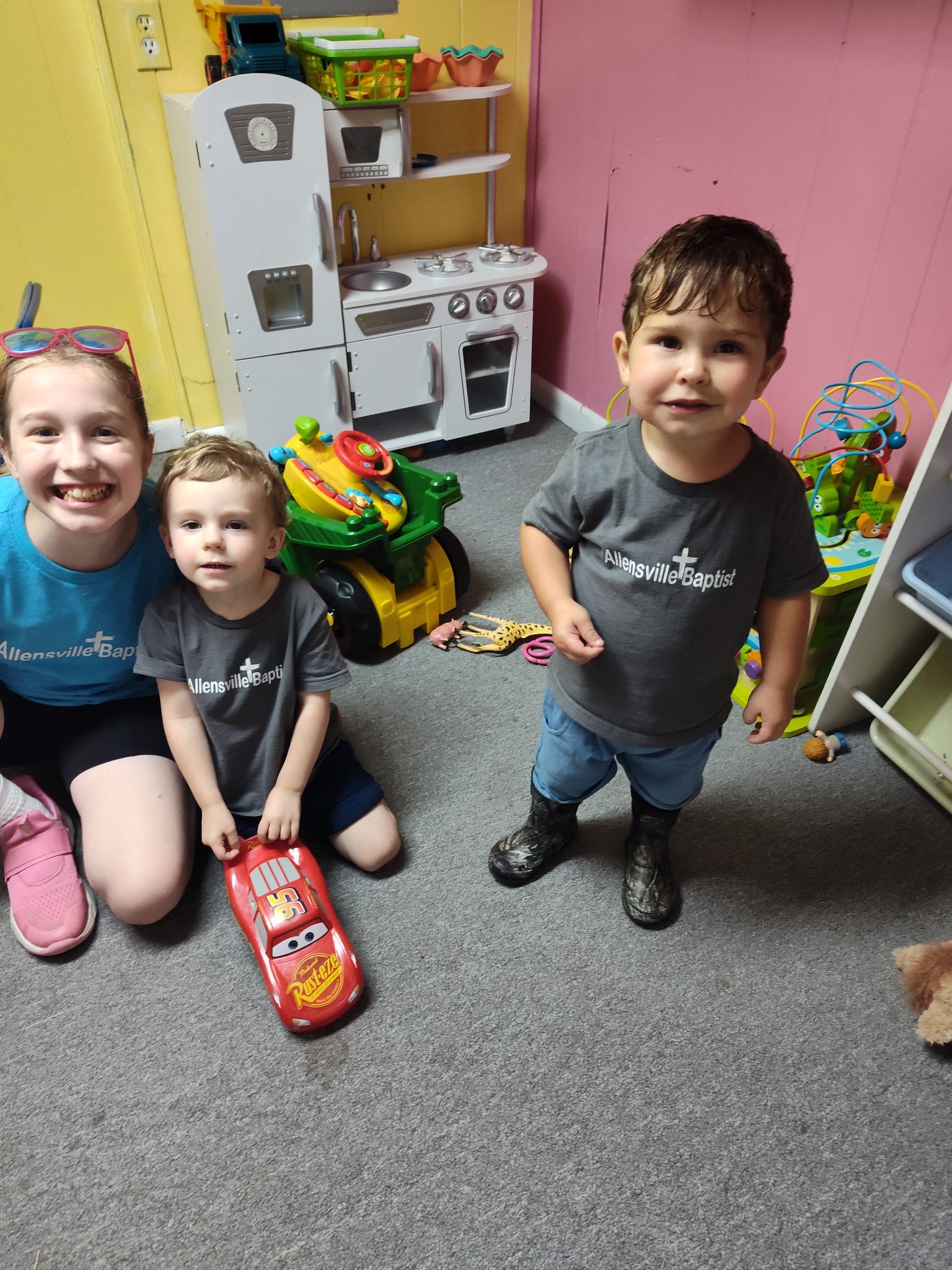 Three children in a playroom, two boys and a girl. The girl is smiling, wearing pink glasses on her head, and sitting on the floor with a toy car. One boy is standing, wearing a gray shirt that says 'Allensville Baptist' and black boots, smiling at t