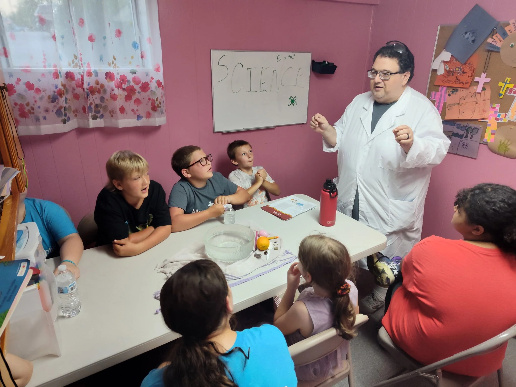 A man in a white lab coat explaining science to a group of children seated around a table in a classroom, with science posters on the wall and a whiteboard with lab equations.