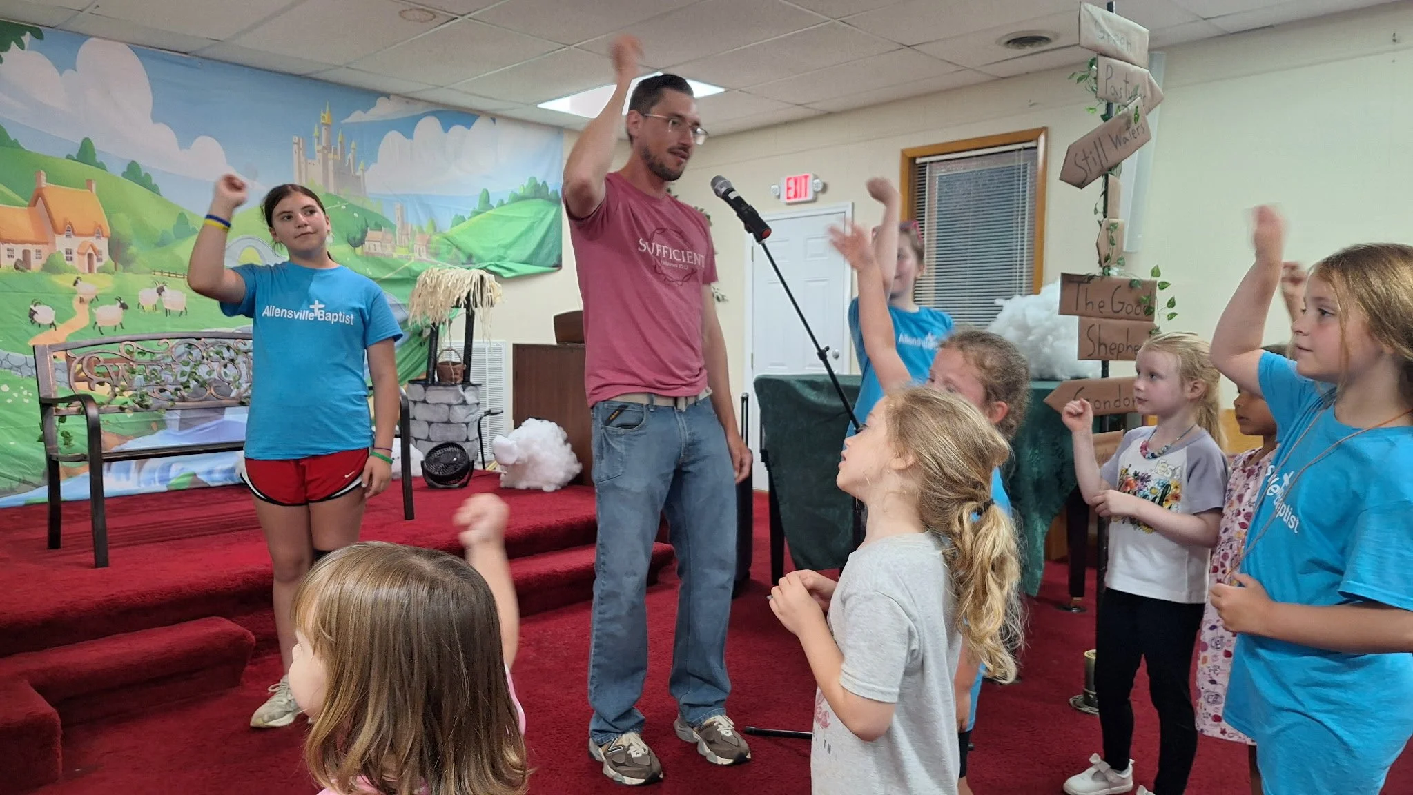 Group of children and a man on stage at a church, with children raising their hands, in front of a colorful backdrop depicting a fantasy castle scene.