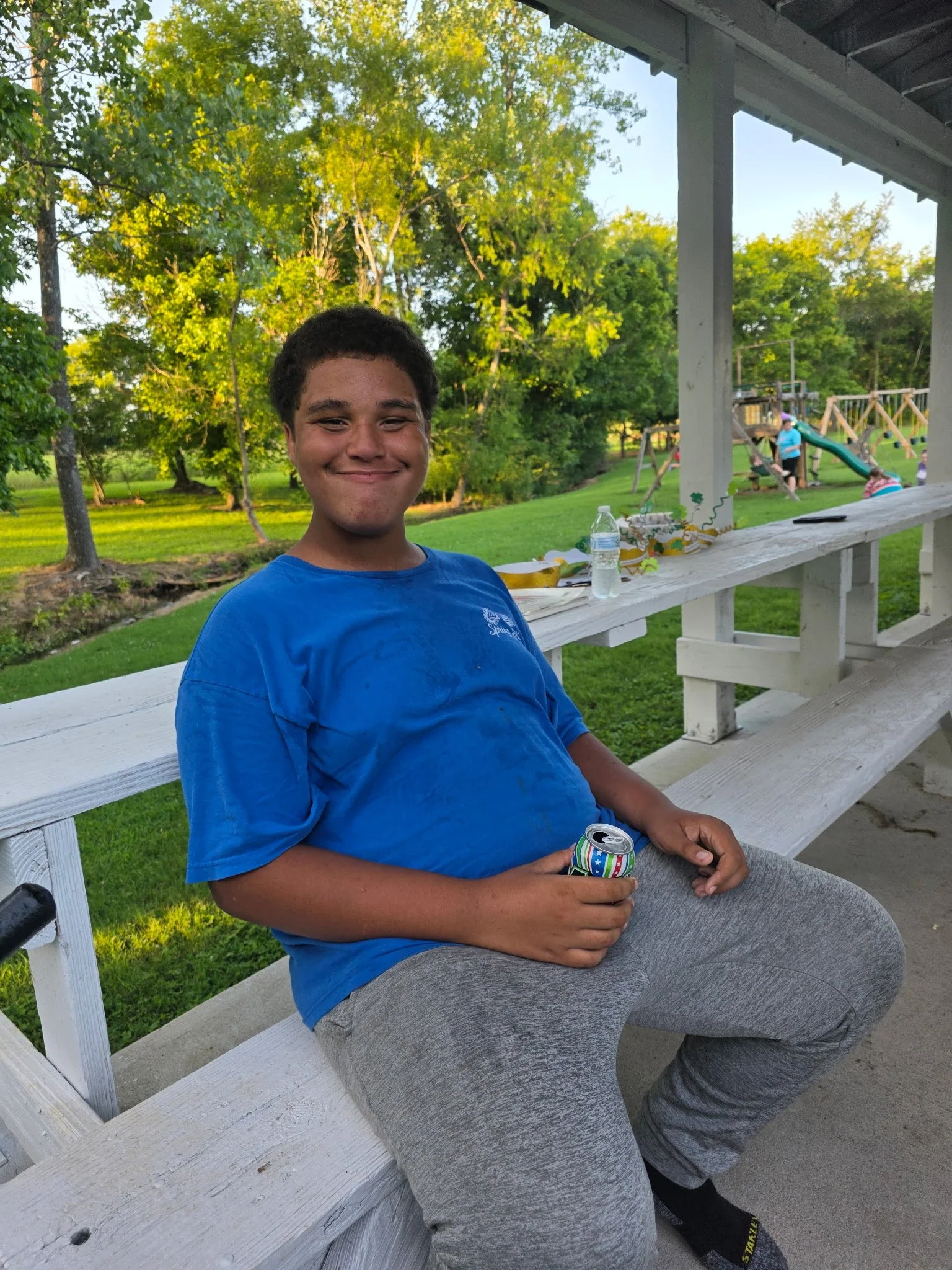 A young man in a blue t-shirt and gray sweatpants sitting on a white wooden picnic table at a park during the daytime. He is smiling and holding a colorful soda can in his hand. In the background, there are big green trees, a grassy area, and a playg