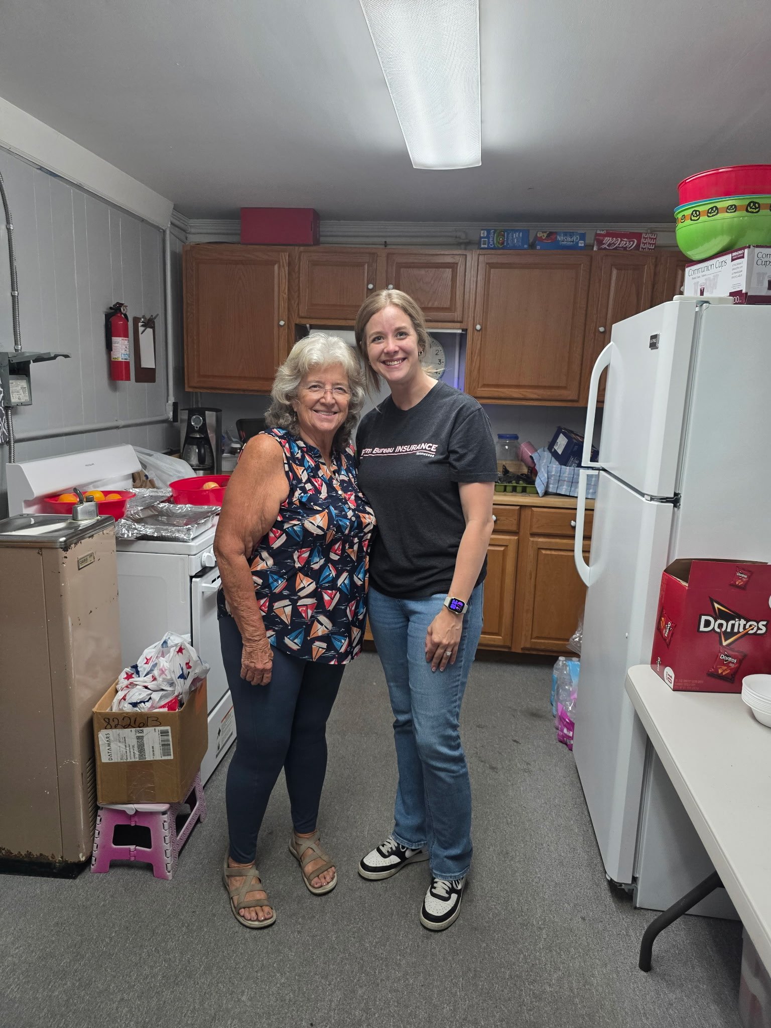 Two women, one older and one younger, standing together in a kitchen, smiling at the camera. The older woman has gray hair and is wearing a colorful sleeveless top and black pants. The younger woman has short brown hair, is wearing a black t-shirt an