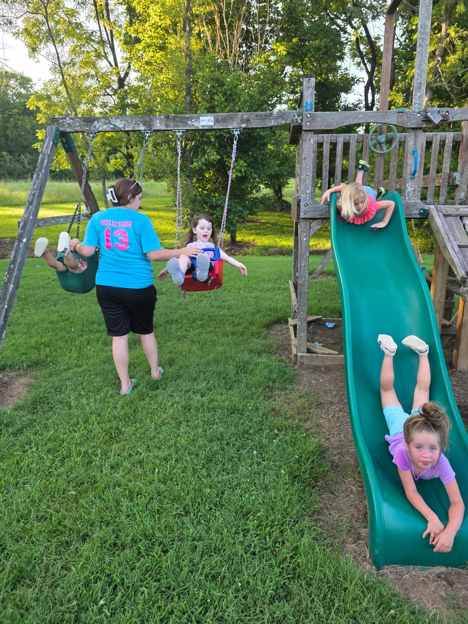 Children playing on a playground swing set and slide outdoors on a grassy area with trees in the background.