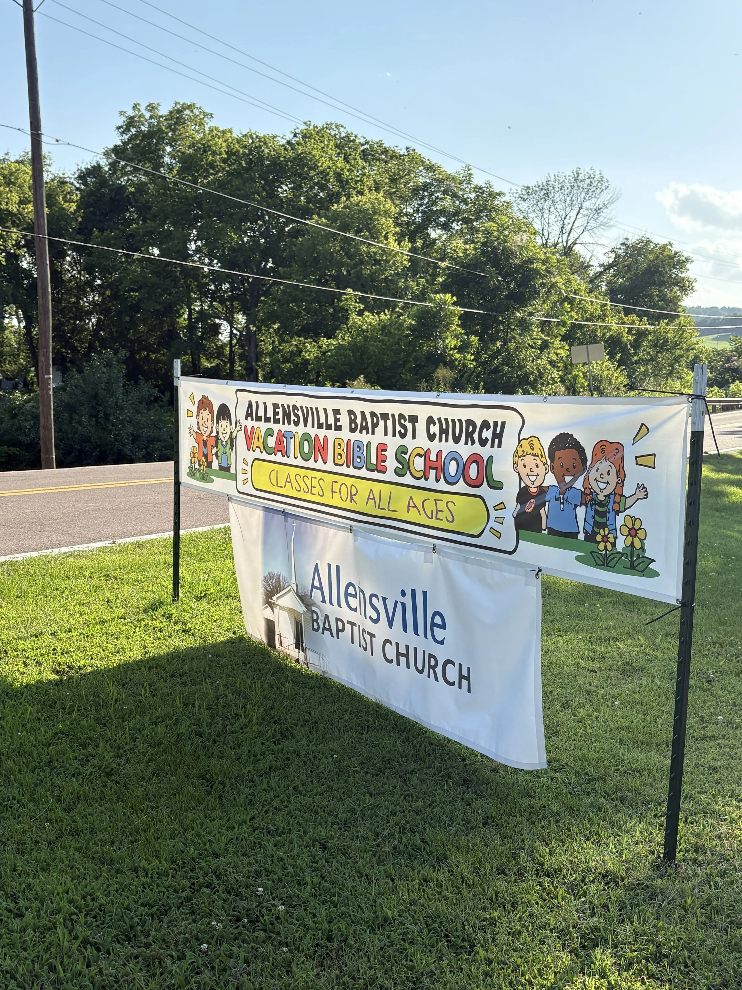 A banner for Allensville Baptist Church Vacation Bible School with colorful text and children's illustrations, placed outdoors on a grassy area beside a road.