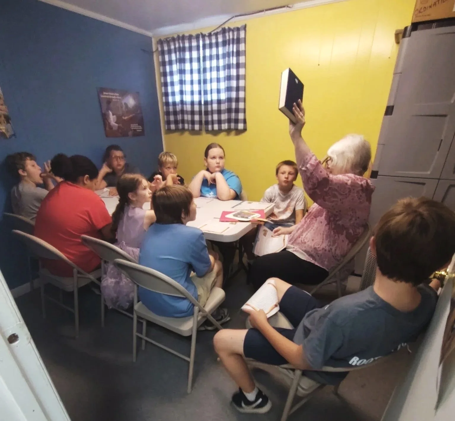 An elderly woman with white hair and glasses is sitting on a beige folding chair, holding a black box up in front of a group of children sitting at a rectangular table in a room with blue and yellow walls. The children are watching her, some with hands on their chins, and appear attentive. There are books and papers on the table, and a window with a checkered blue and white curtain on the yellow wall.