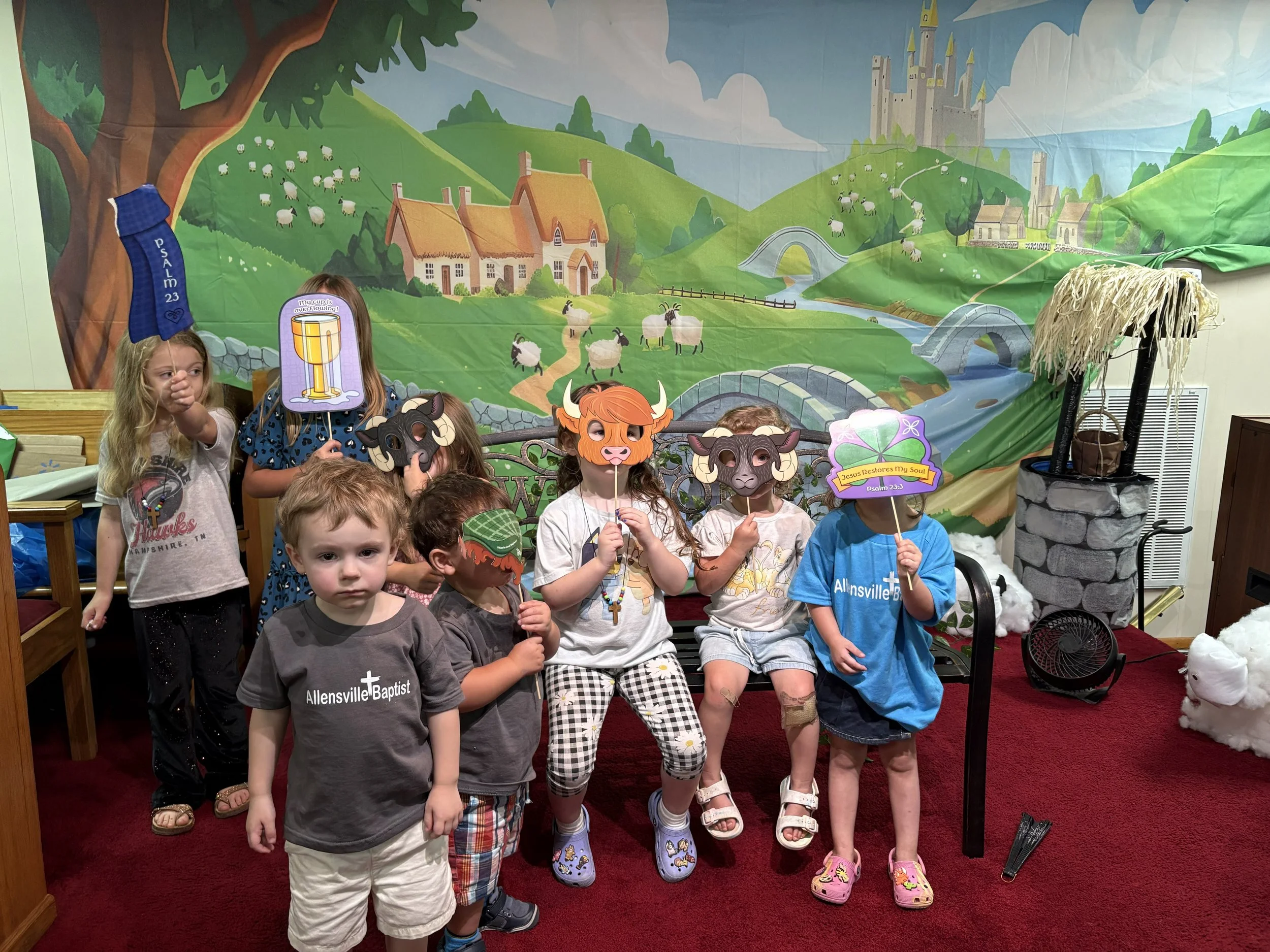 Children participating in Vacation Bible School, with some holding animal masks and signs, standing in front of a colorful mural depicting a farm with sheep, a castle, and a river.