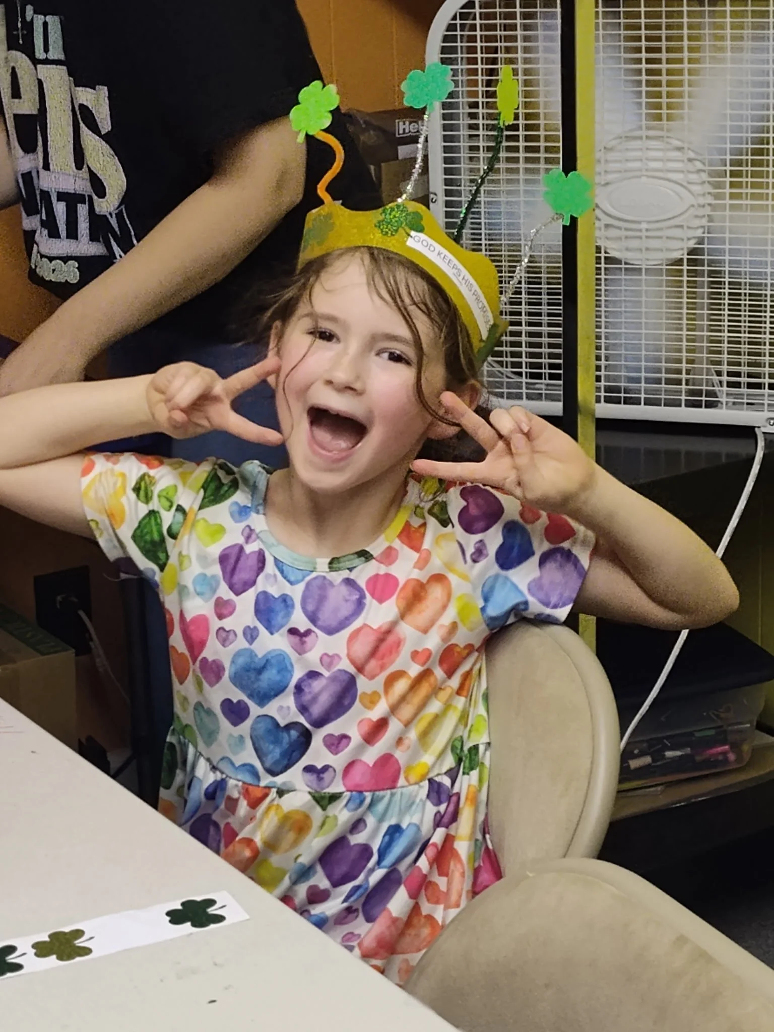 A girl wearing a rainbow-colored heart patterned dress and a yellow St. Patrick's Day hat with shamrocks and the words 'God keeps his promises' is sitting in a beige chair, making a peace sign with both hands near her face, smiling widely, in front o