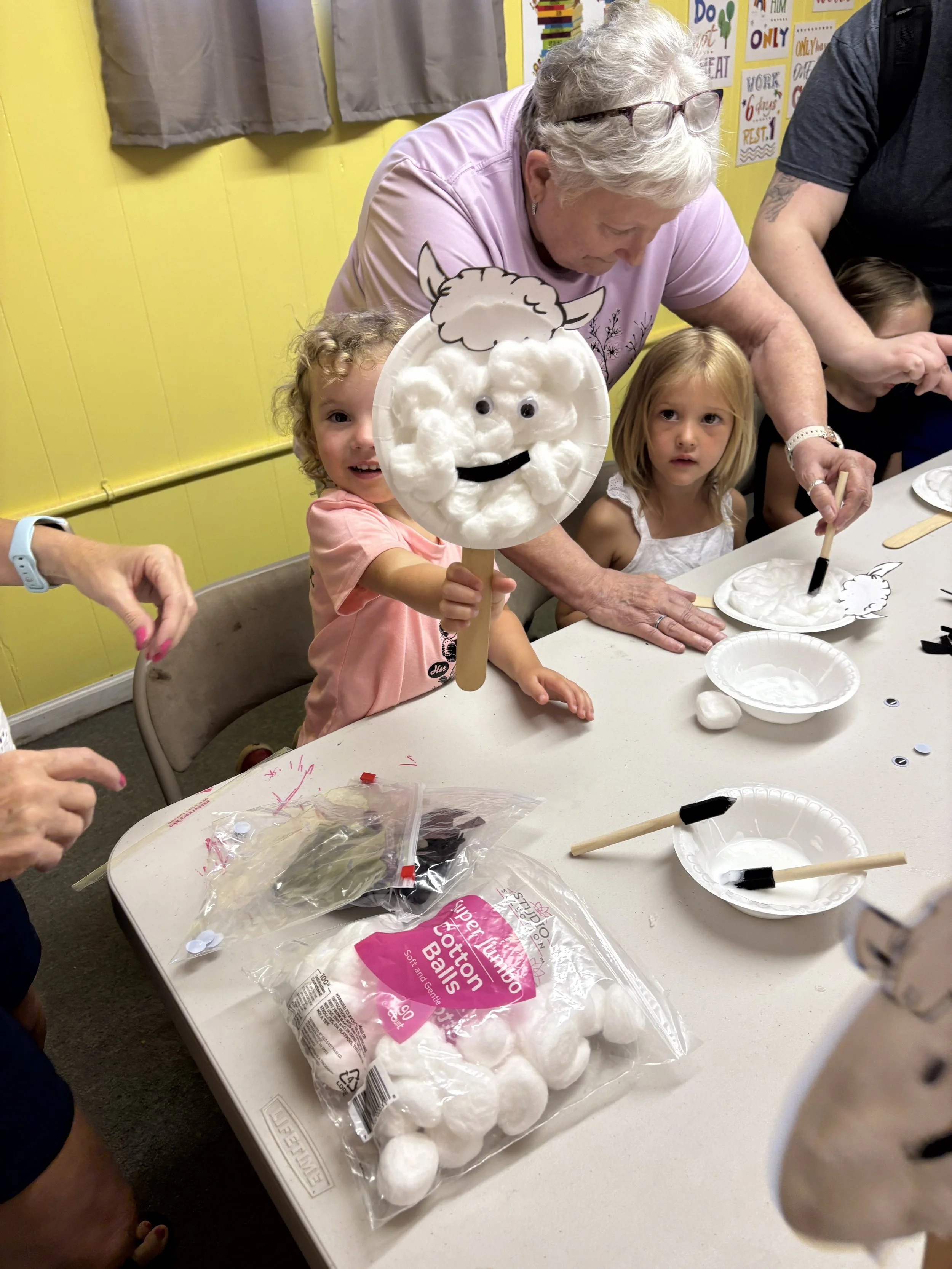 A smiling young girl holding a cotton ball craft with a sheep's face, alongside an older woman and another girl at a table with craft supplies in a classroom.