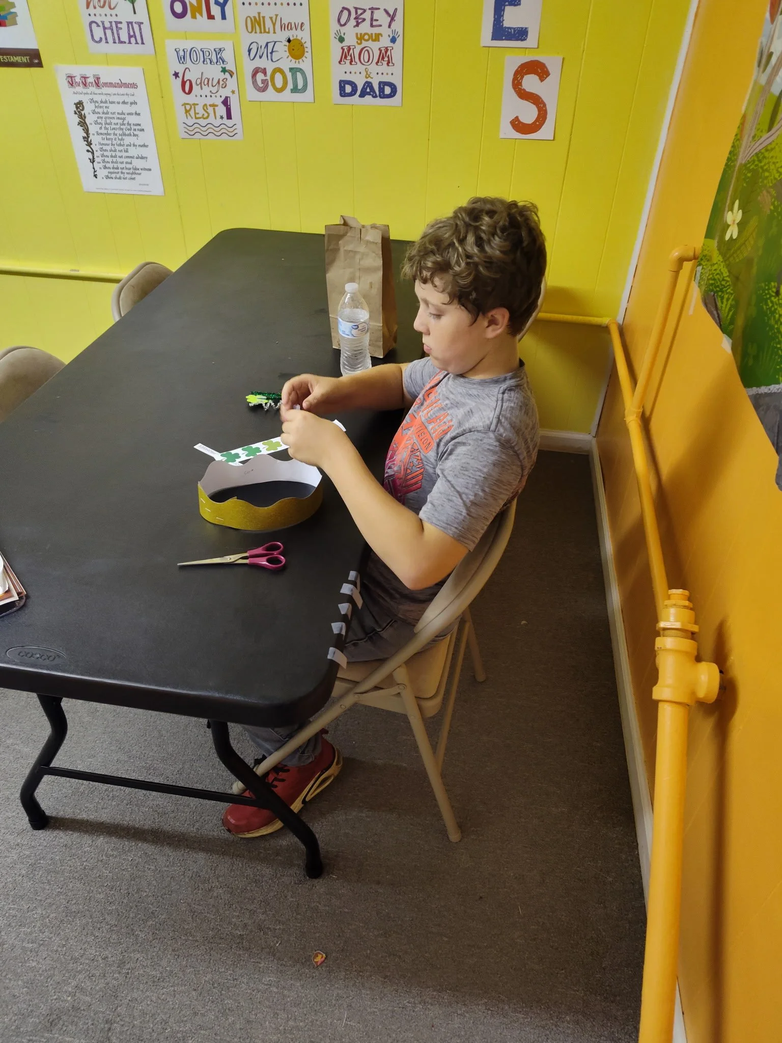 A young boy with curly brown hair wearing a gray T-shirt and red shoes sitting at a black table. On the table are craft supplies, including scissors, a paper crown, and stickers. Behind him is a yellow wall decorated with posters and signs, including