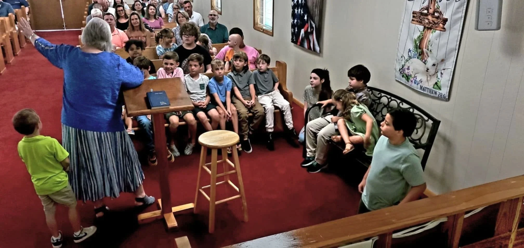 A woman in a blue top and striped skirt is speaking to a group of children and adults in a church or community room. The children are seated on pews and some are standing around, listening attentively.