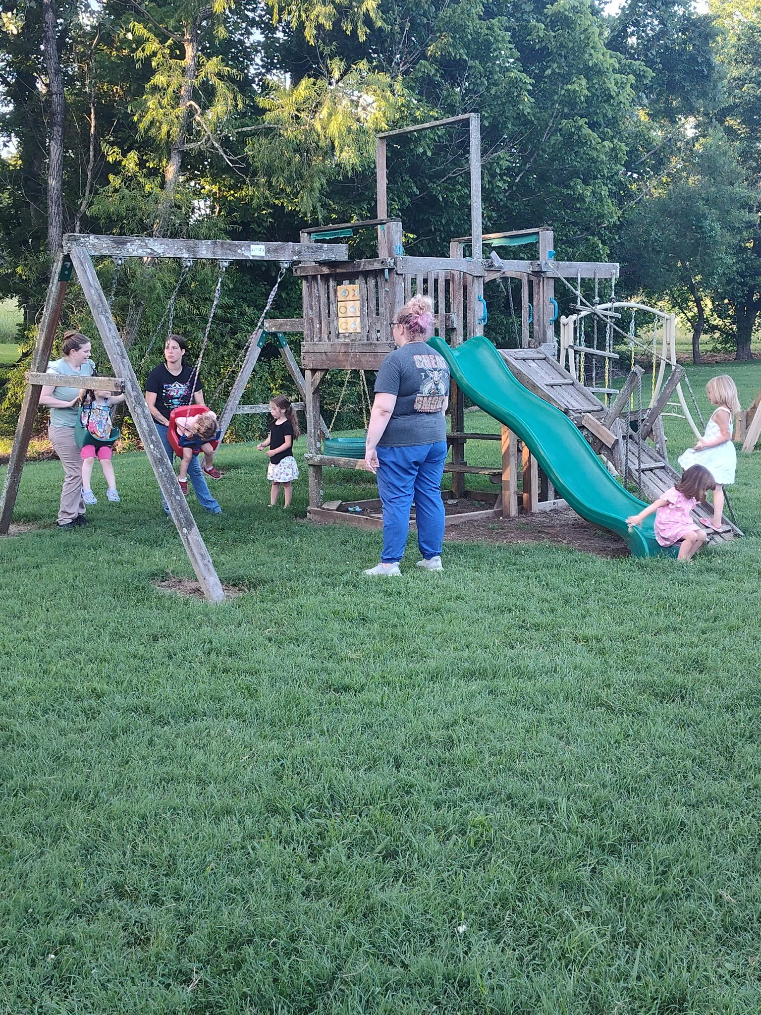 A playground with a wooden play structure, swings, and a slide, surrounded by grass and trees, with children and adults playing and supervising.