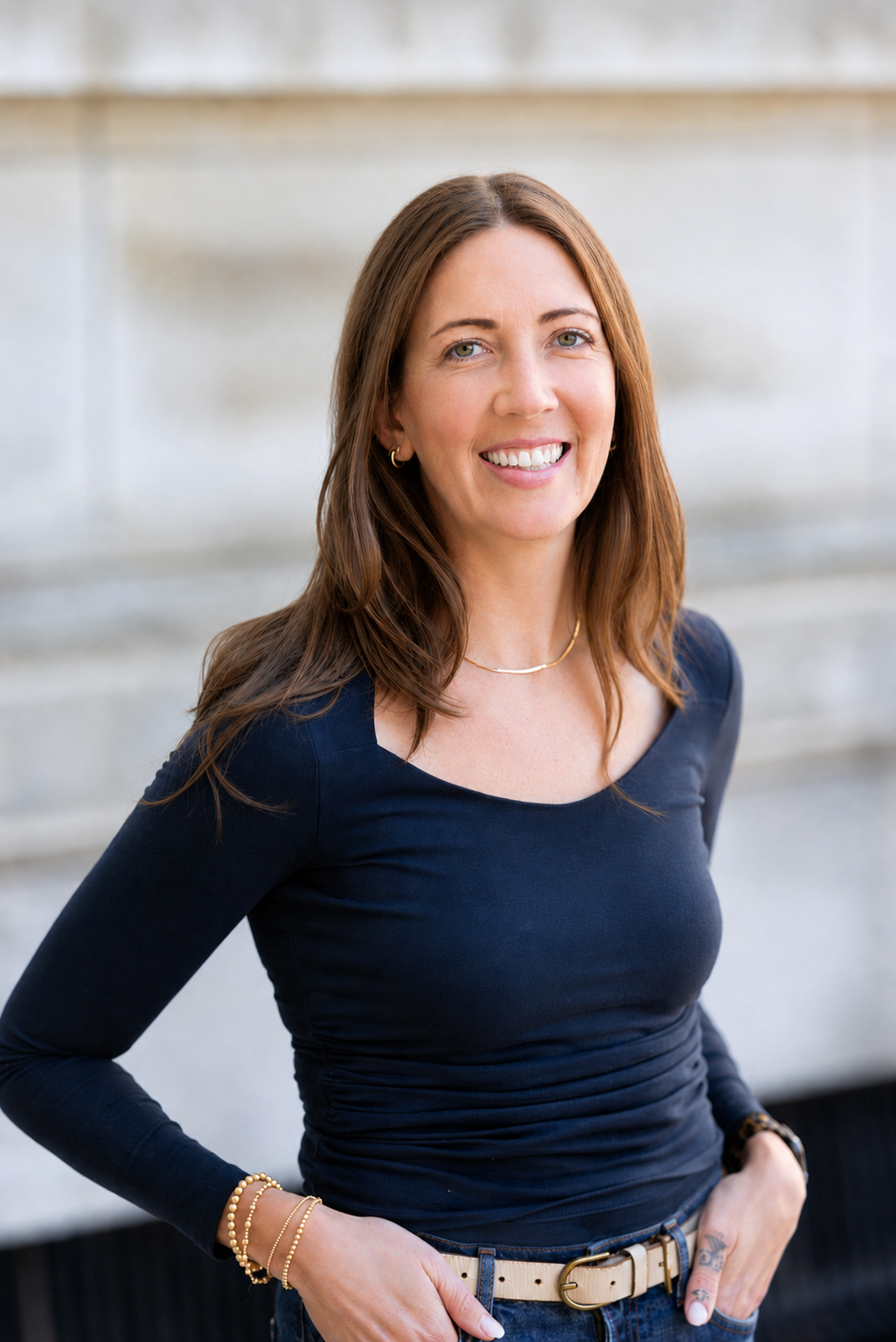 A young woman with long, light brown hair, smiling and wearing a white top, standing against a plain wall background.