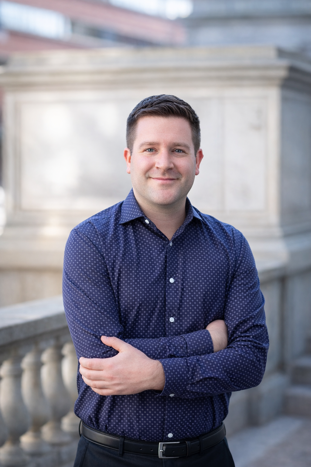 Portrait of a man with short brown hair, blue eyes, and light skin, smiling slightly, wearing a dark blue patterned button-up shirt, outdoors with blurred background.