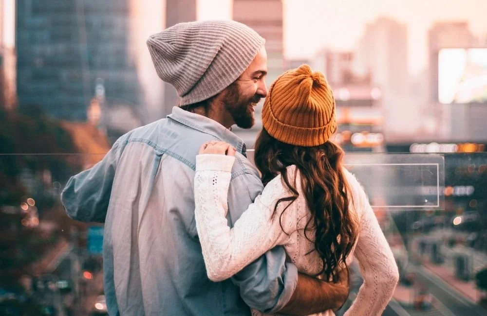 A couple with beanies on standing on a bridge overlooking the city while the man smiles down at the woman holding her close | Kathleen Killen Psychotherapy and Coaching