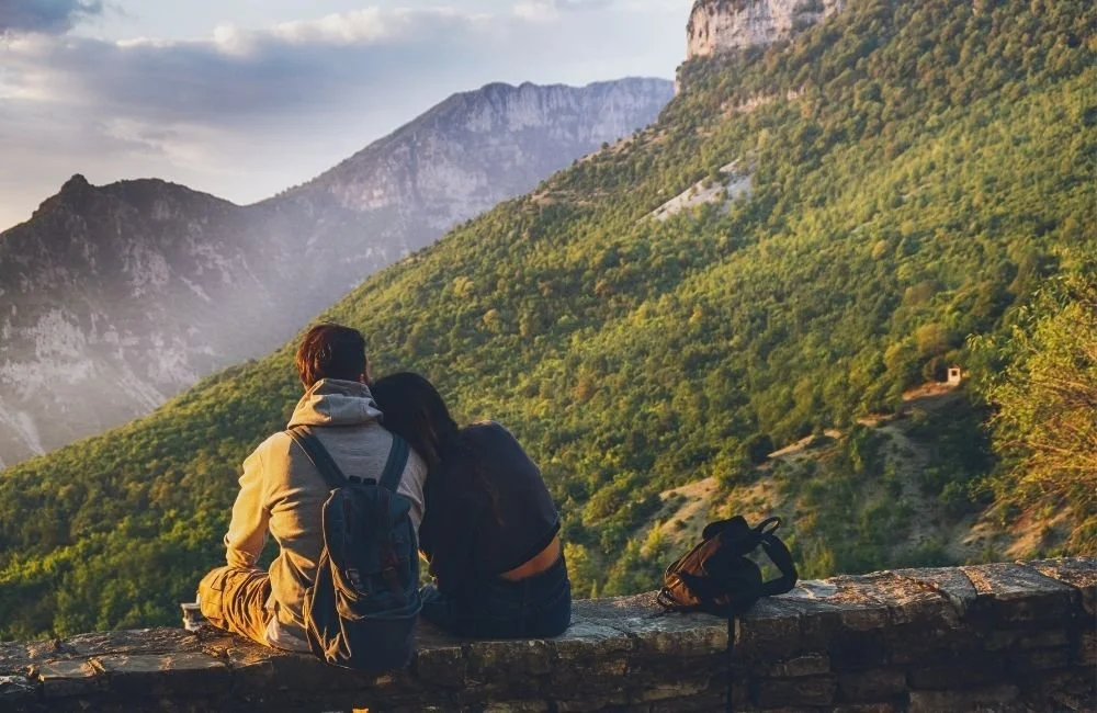 A couple sit on top of a wall overlooking hills and mountains. Find couples travel tips to reduce stress while travelling with your partner and avoid relationship conflict on vacation.